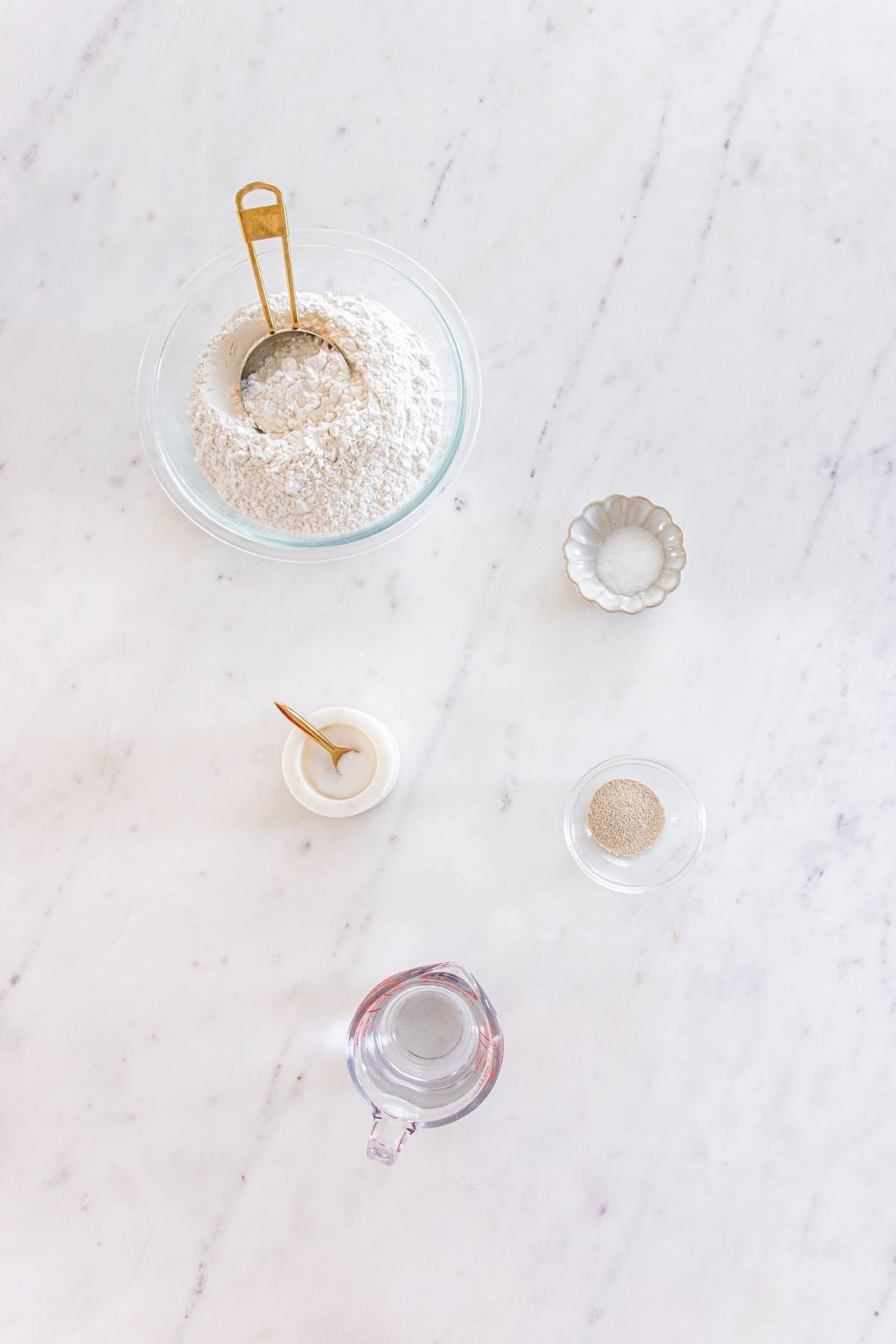 Bowl of flour with a scoop, small bowls of yeast and salt, a cup of water, and a jar on a white surface.