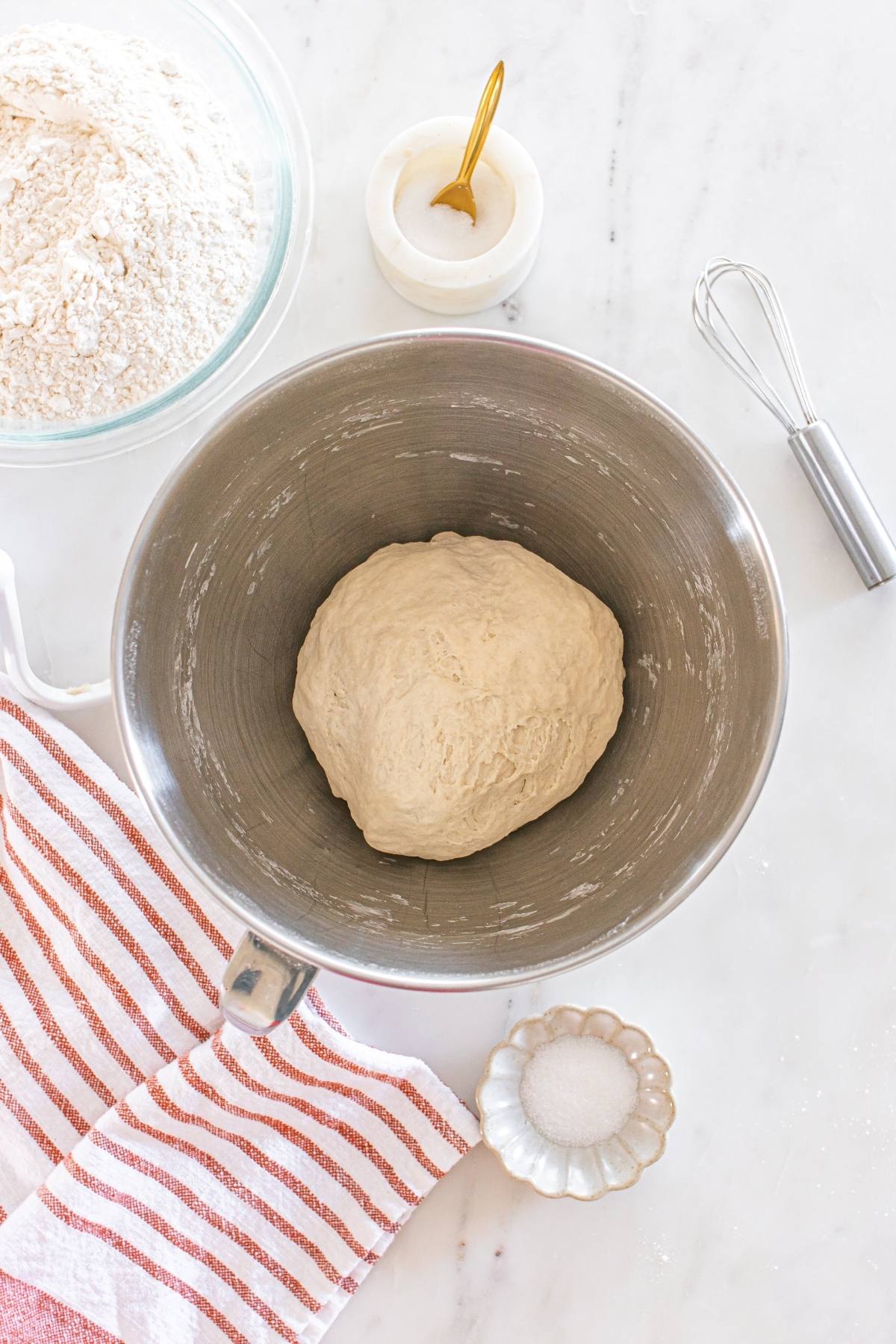 A ball of dough in a metal bowl, with flour, salt, a whisk, and a striped towel nearby on a white surface.