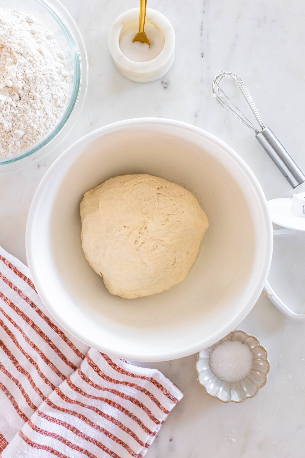 A ball of dough in a white bowl on a marble counter with flour, salt, and a red-striped towel nearby.