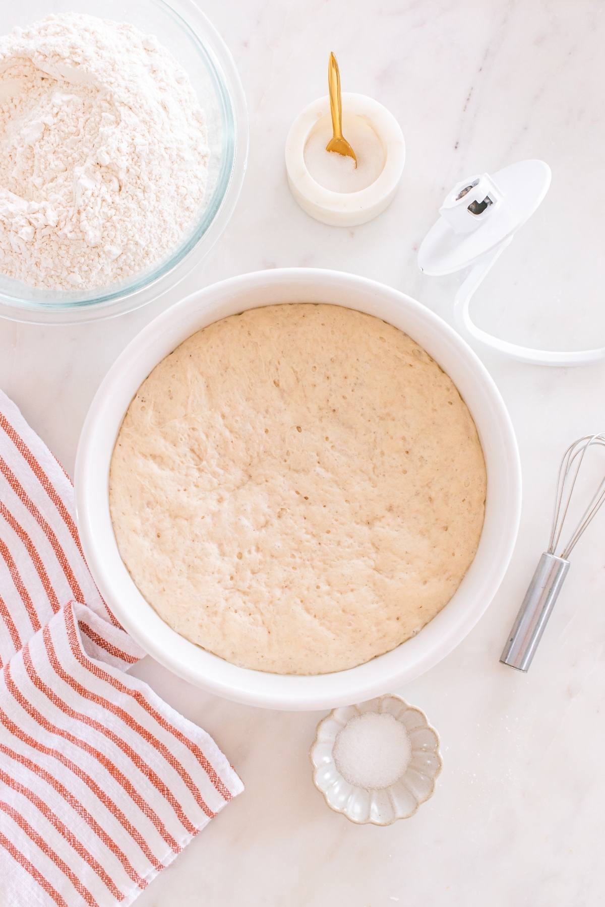 A bowl of risen dough on a marble surface with flour, salt, a whisk, a towel, and a candle nearby.