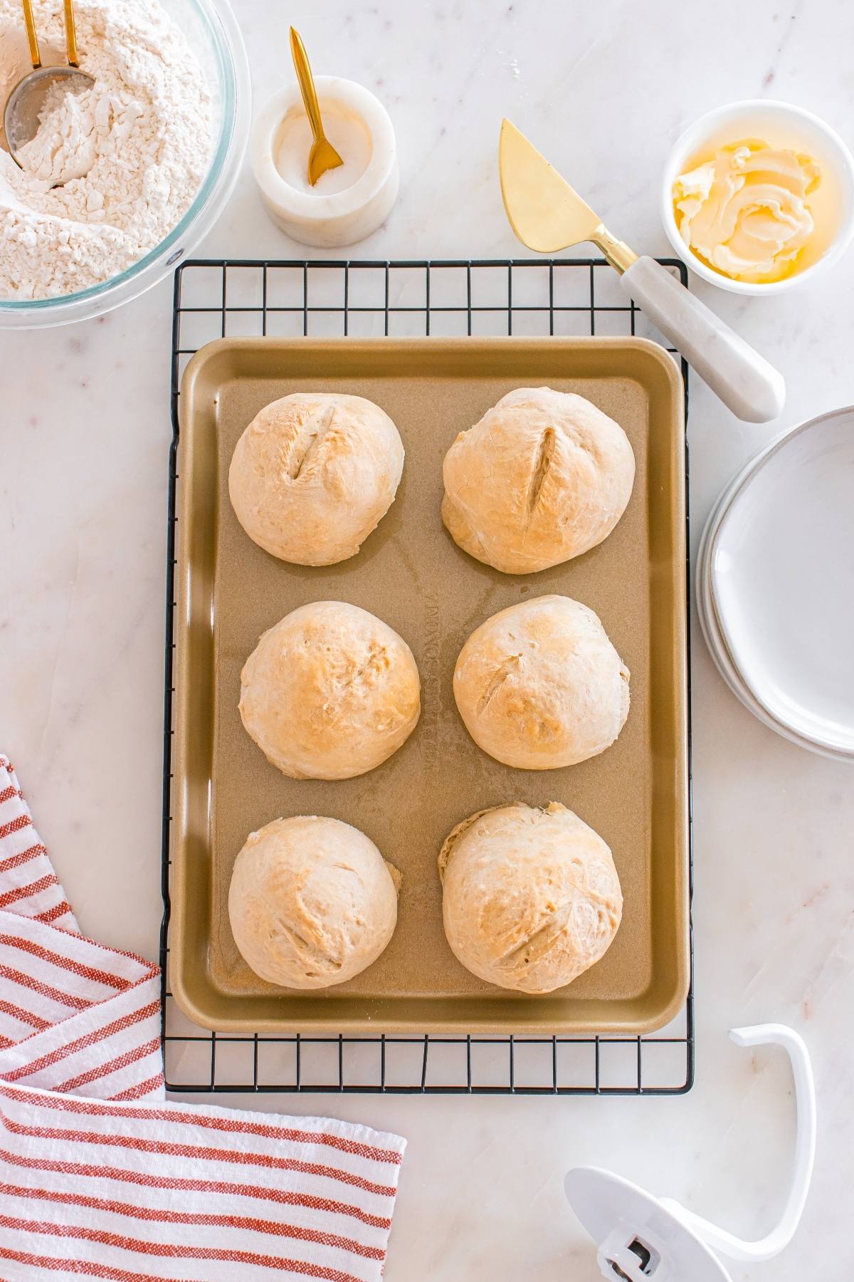 Six baked bread rolls on a gold baking sheet, surrounded by flour, butter, plates, and kitchen utensils.