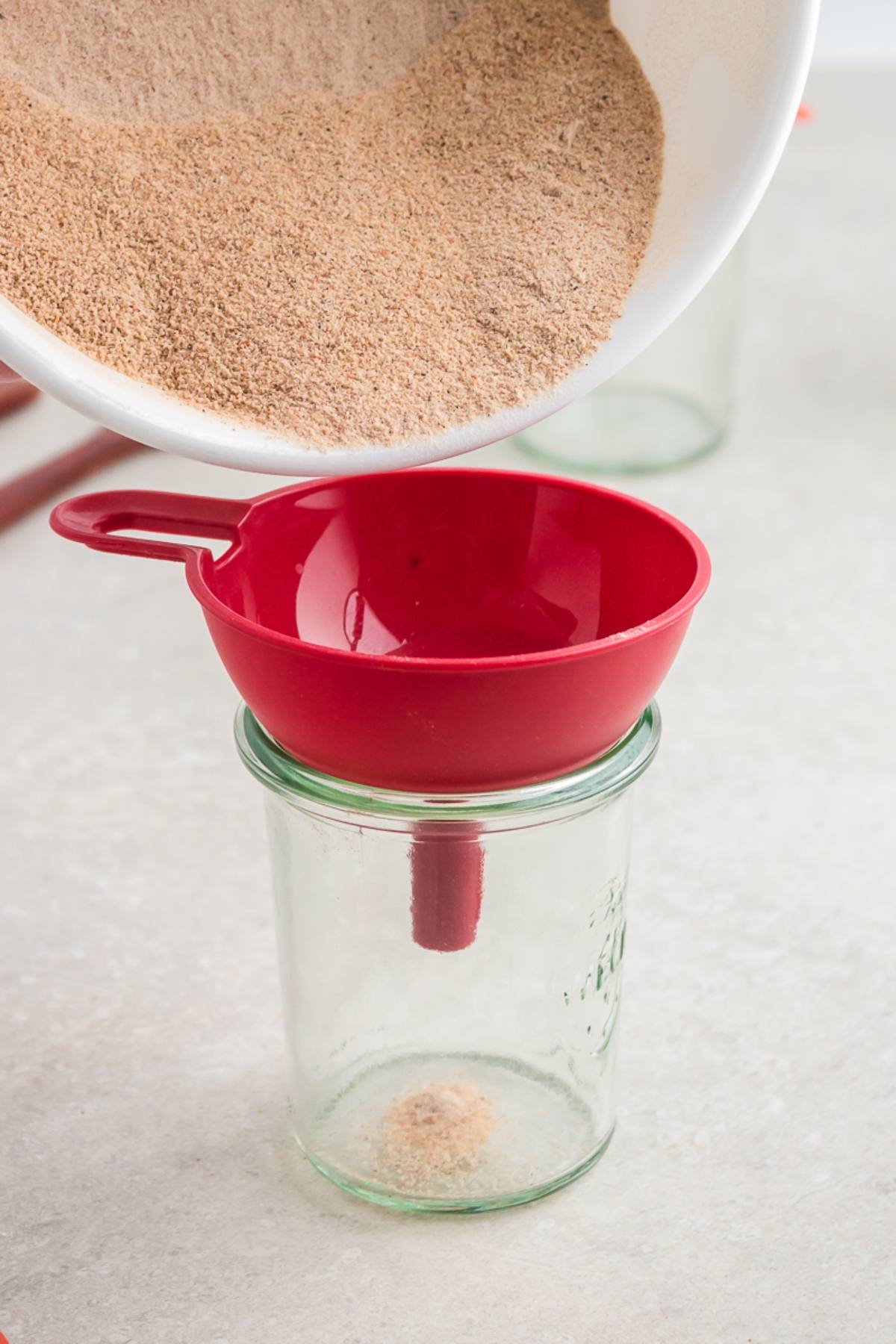 A bowl pours brown powder, perfect for a DIY BBQ Rub Recipe, through a red funnel into an empty glass jar on a light surface.