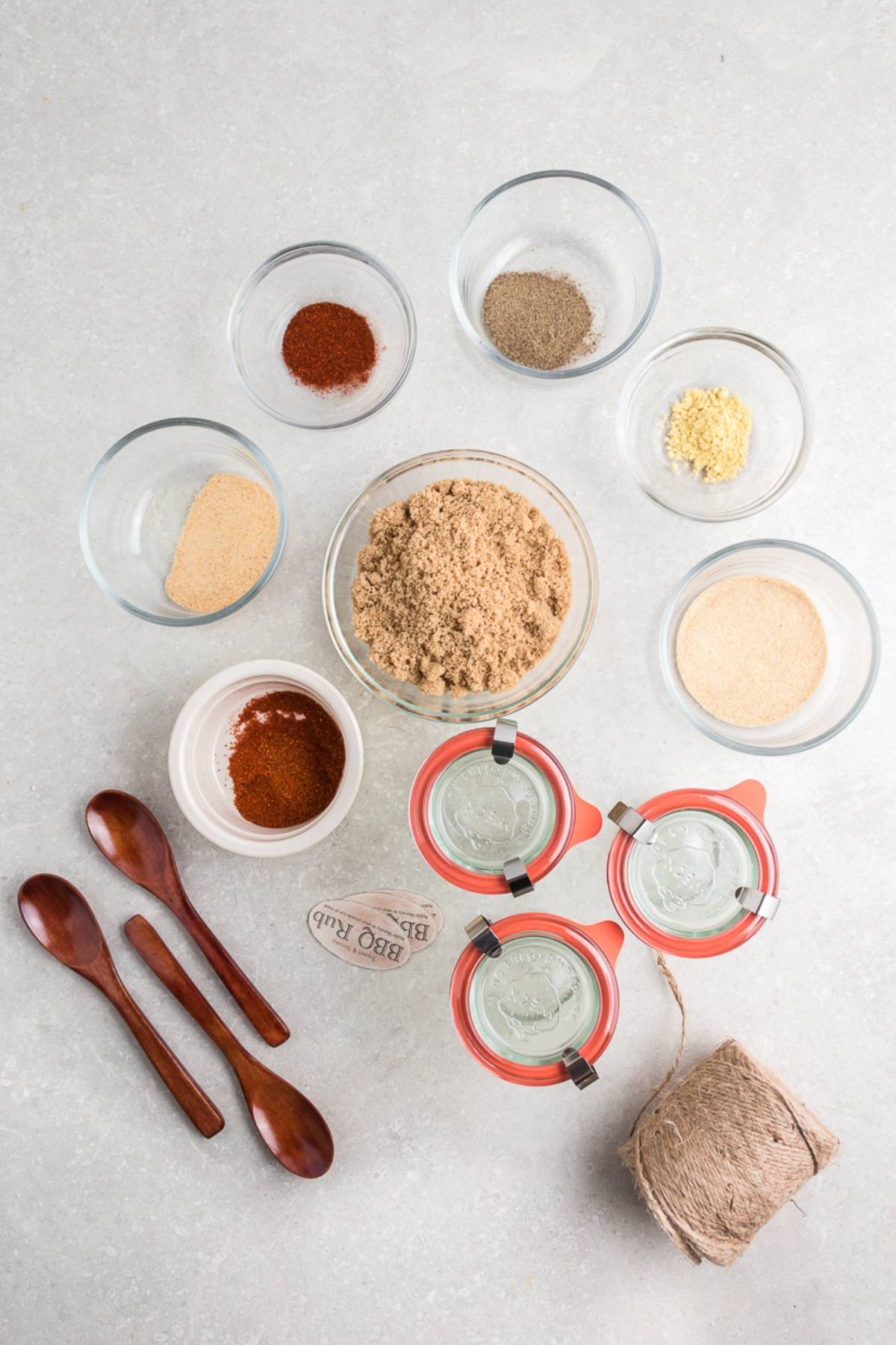 Jars, spices in bowls, wooden spoons, and twine arranged on a light surface for a DIY BBQ Rub Recipe preparation.