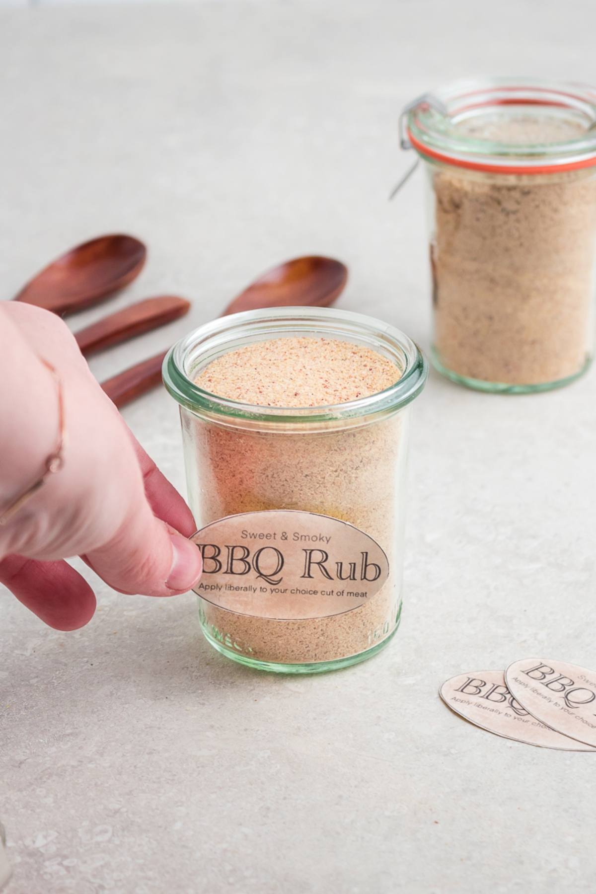 A hand holding a jar labeled "BBQ Rub" on a countertop, surrounded by wooden spoons and another jar—perfect inspiration for your next DIY BBQ Rub Recipe.