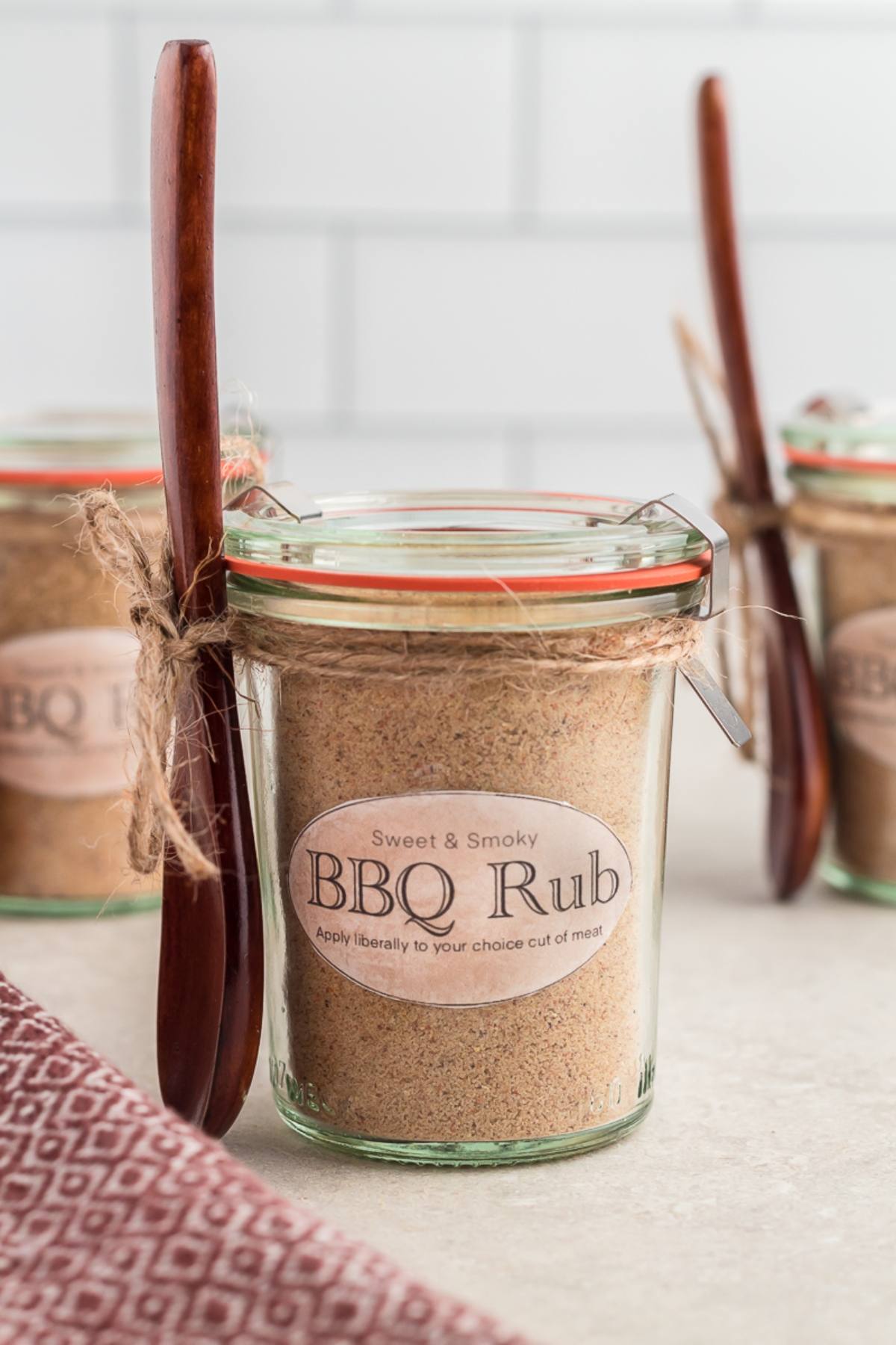 Glass jar labeled "BBQ Rub" with a wooden spoon tied to it, filled with spice mix from a DIY BBQ Rub Recipe, on a countertop.