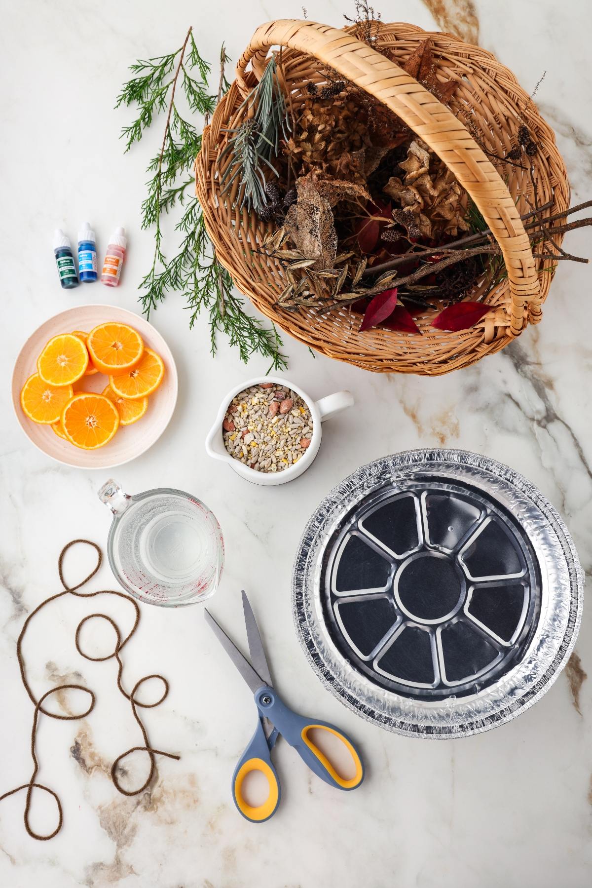 Craft supplies on a table: basket of leaves, orange slices, twine, scissors, foil pan, water, birdseed, food coloring.