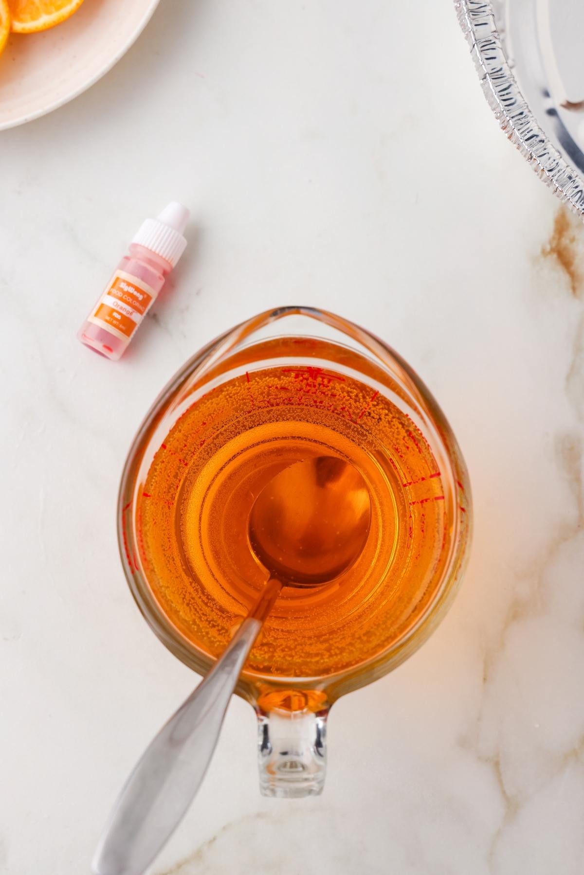 A glass measuring cup with orange liquid and a spoon, next to a small bottle of orange food coloring.