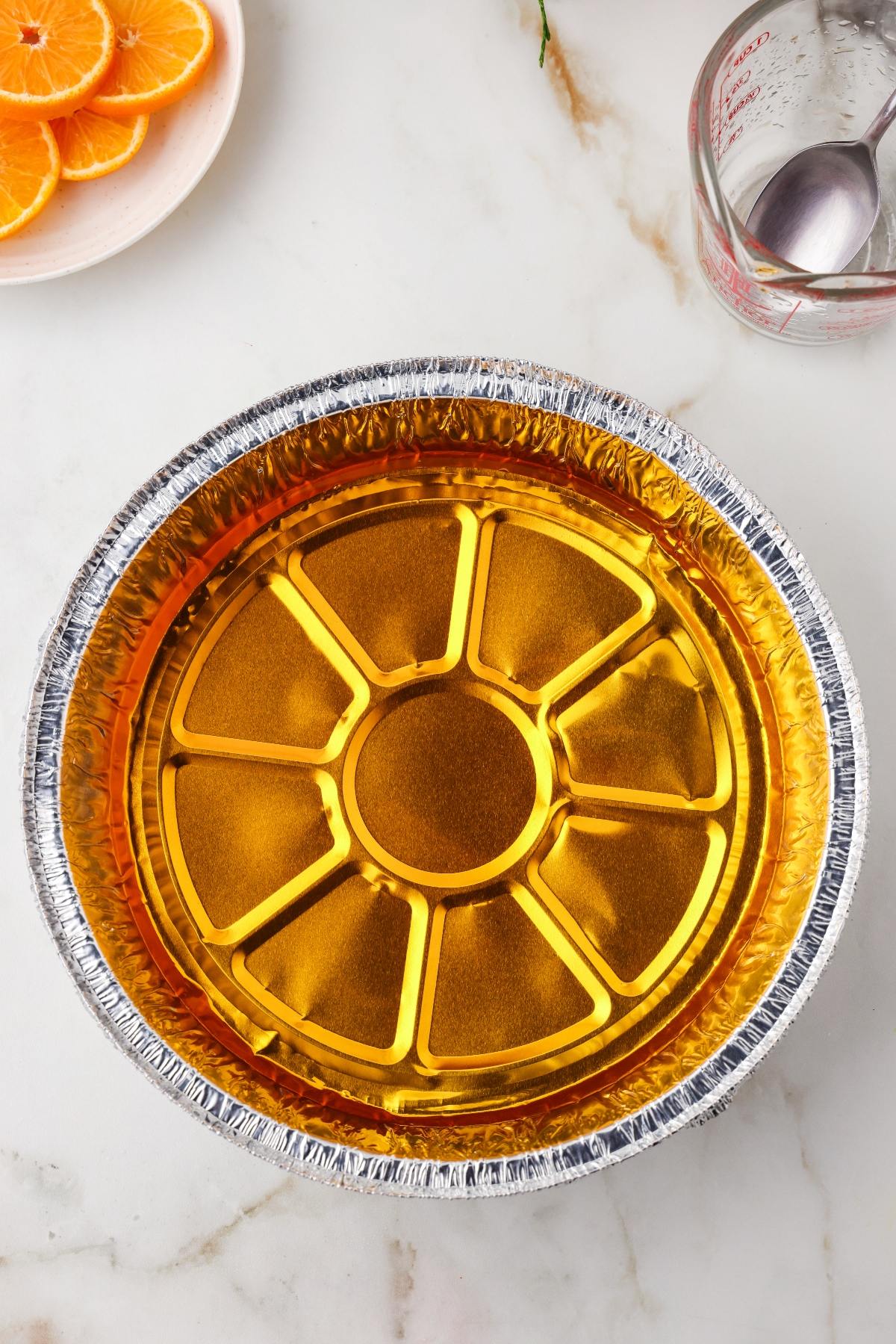 A round aluminum pan filled with orange gelatin sits on a marble surface next to orange slices and a measuring cup.