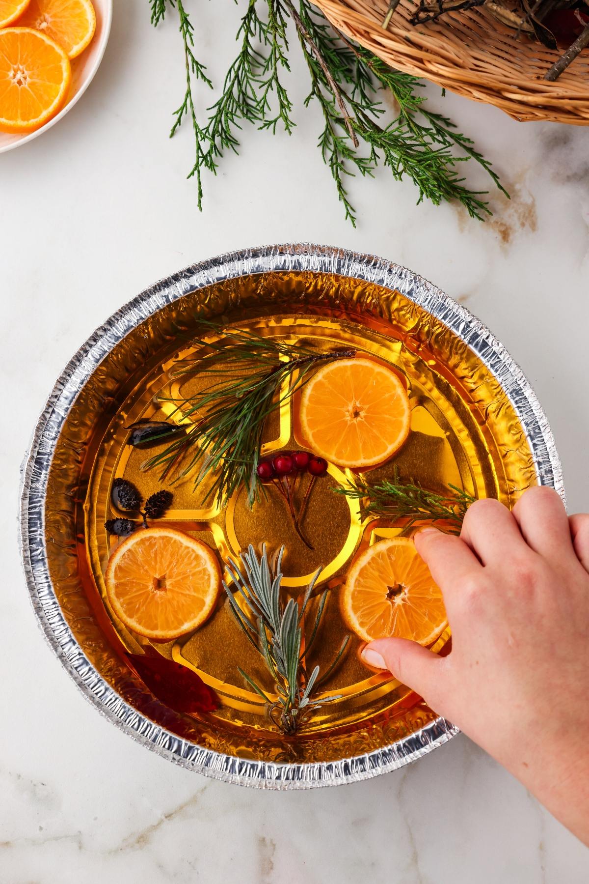 A hand arranges orange slices and herbs in a round foil pan filled with liquid on a white surface.