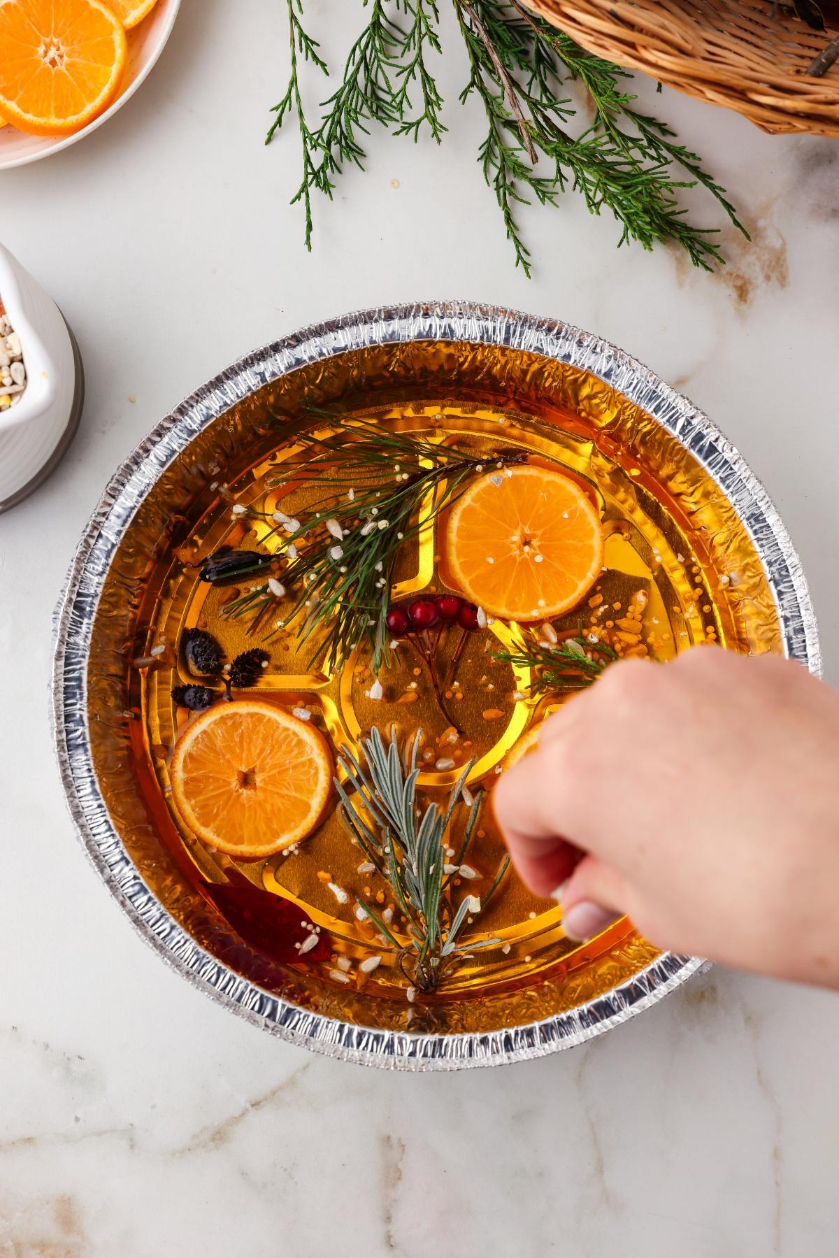 A hand sprinkles seasoning into a pan of simmering potpourri with orange slices and rosemary.