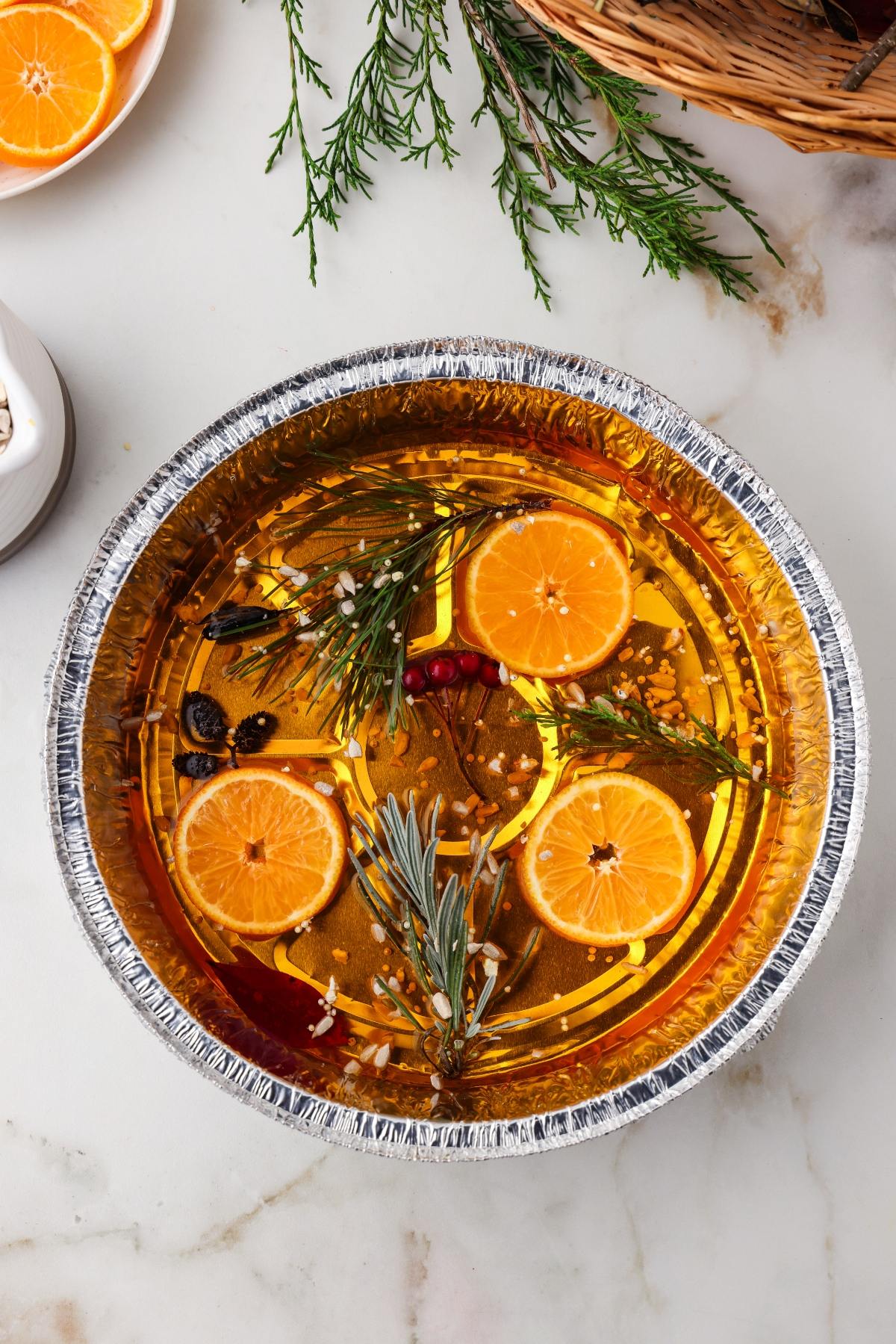 A foil pan filled with sliced oranges, herbs, spices, and liquid, arranged on a marble countertop.