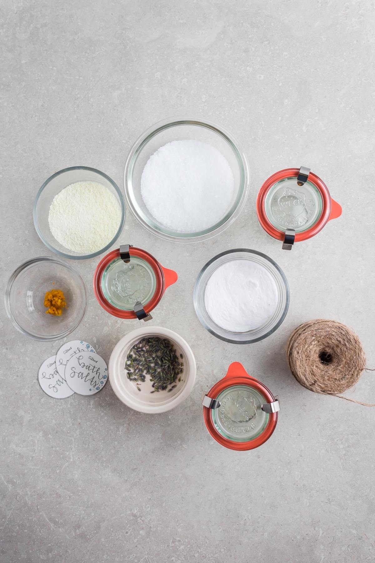 Top view of jars and bowls with DIY Lavender Orange Bath Salts ingredients, twine, tags, and dried lavender on a gray surface.