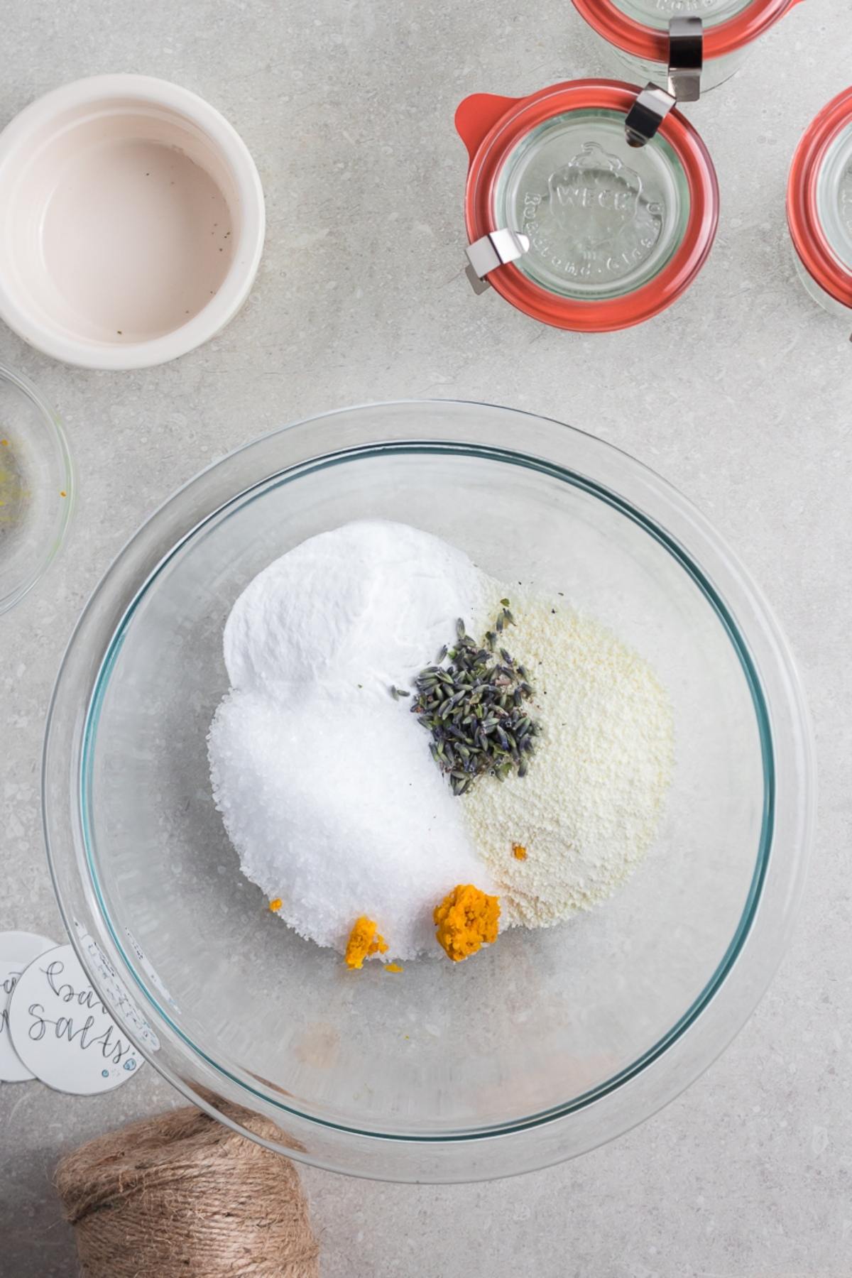 A glass bowl with salt, lavender, and yellow powder sits on a gray counter, surrounded by jars and twine—perfect for making your own DIY Lavender Orange Bath Salts.