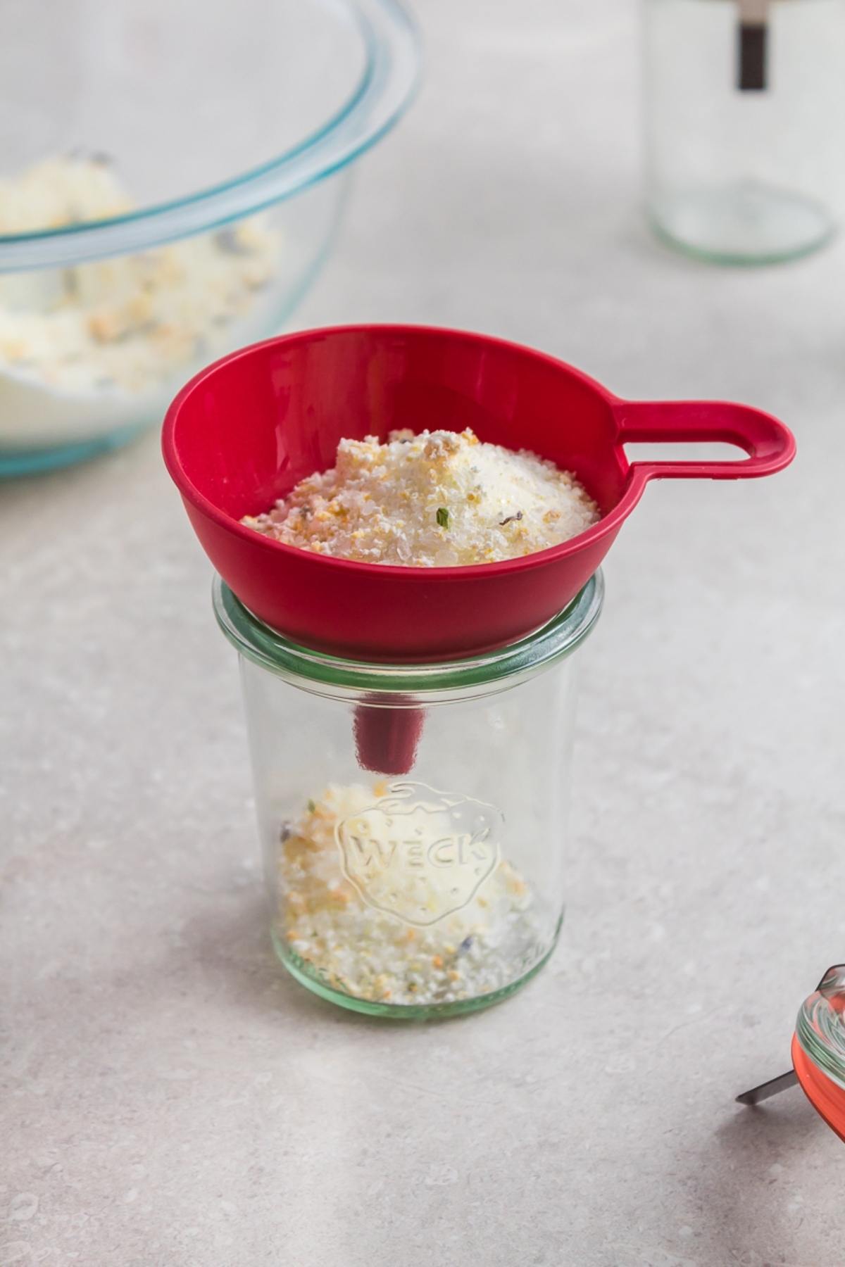 A red funnel pours a crumbly mixture of DIY Lavender Orange Bath Salts into a glass jar on a light countertop.