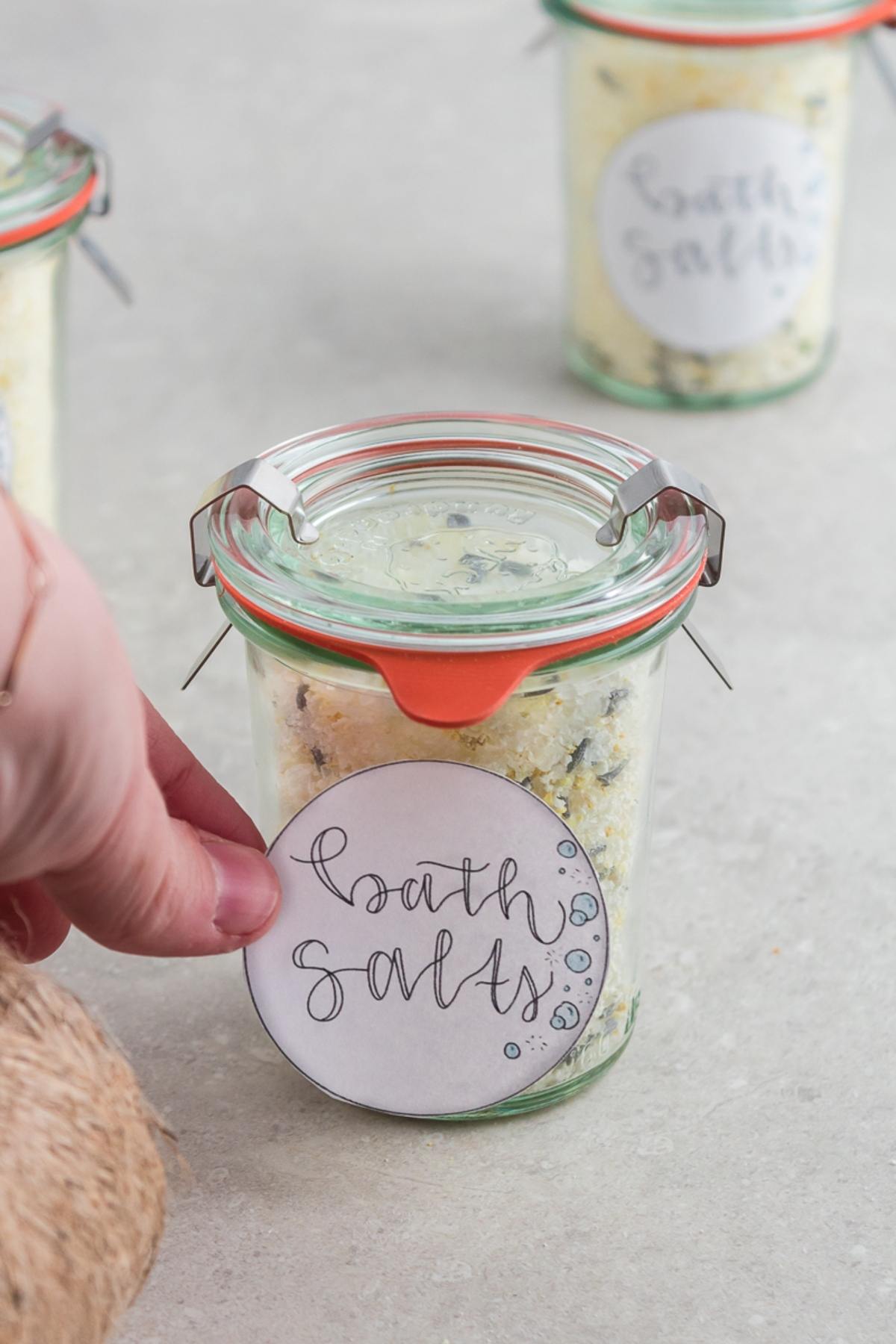 A hand holds a jar labeled "bath salts" filled with homemade DIY Lavender Orange Bath Salts on a light surface.