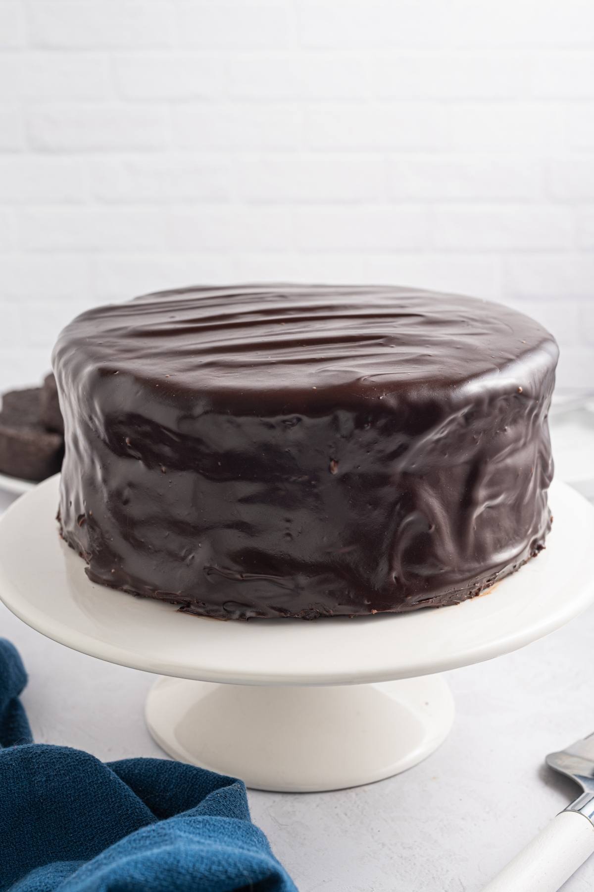 A round chocolate cake covered in glossy chocolate ganache on a white cake stand.