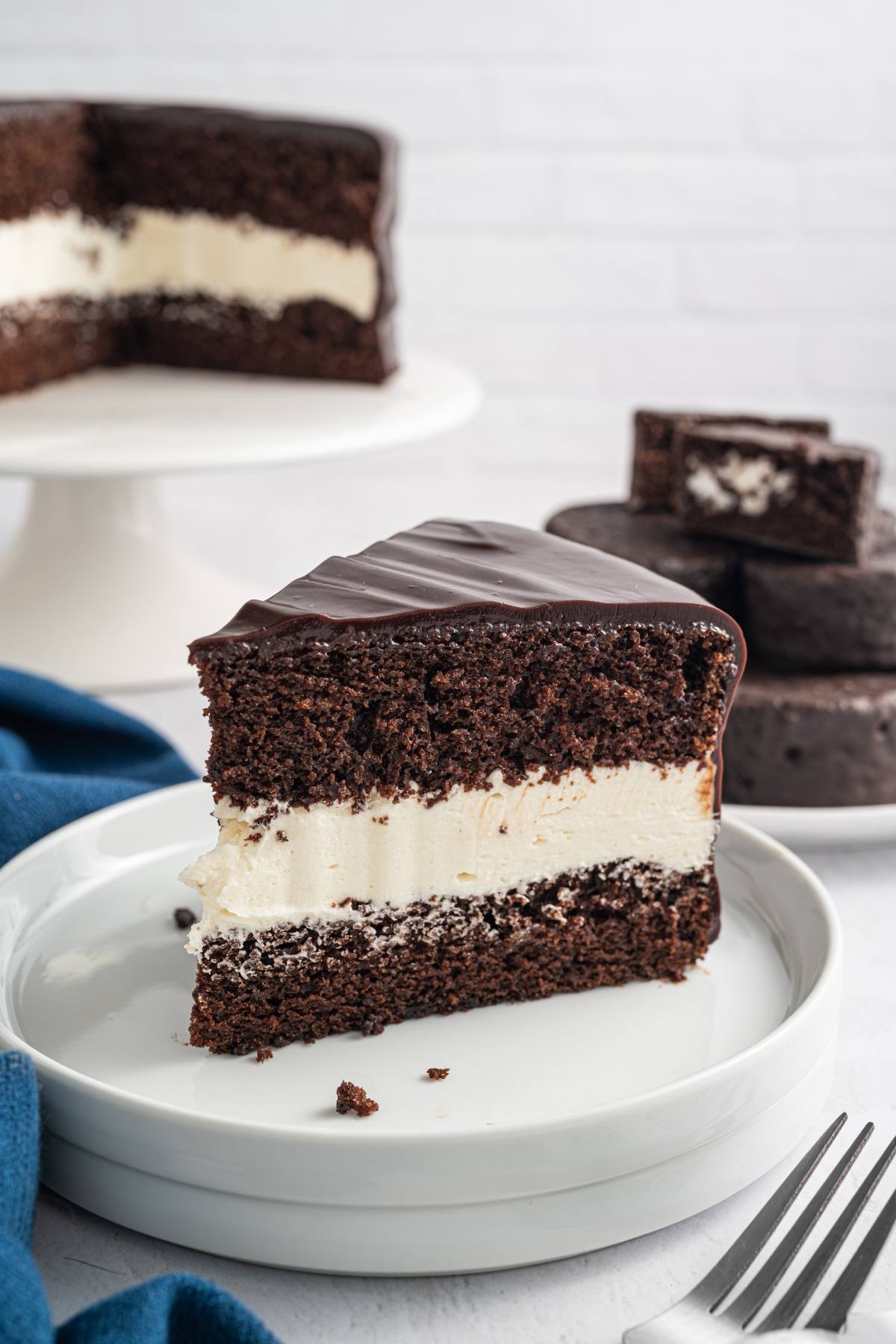 A slice of chocolate cake with cream filling on a plate, with the rest of the cake in the background.