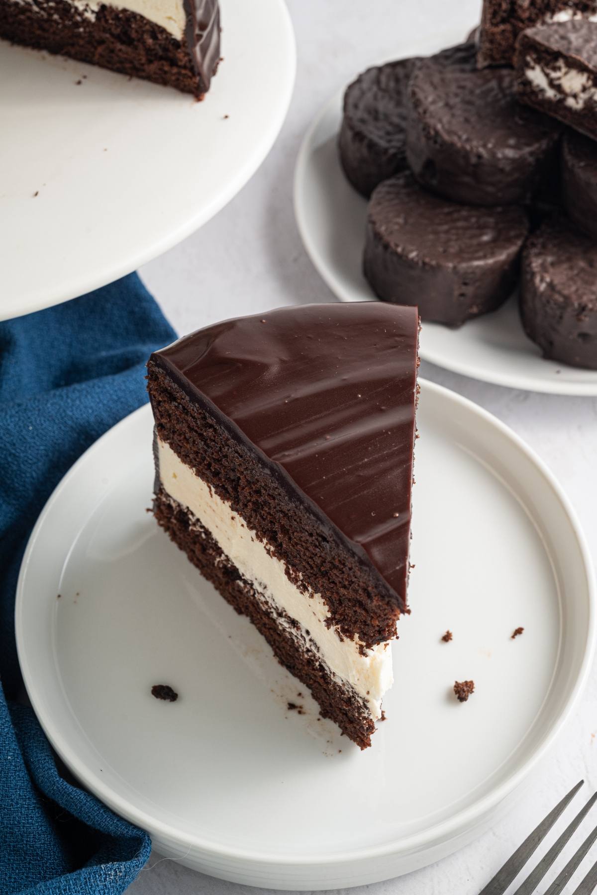 A slice of chocolate cake with cream filling on a white plate, near a stack of chocolate-covered cakes.