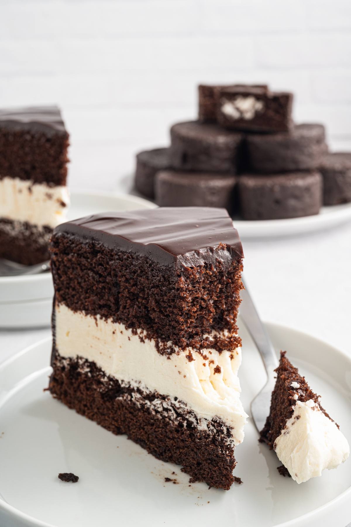 A slice of chocolate layer cake with cream filling and glossy chocolate icing on a white plate.