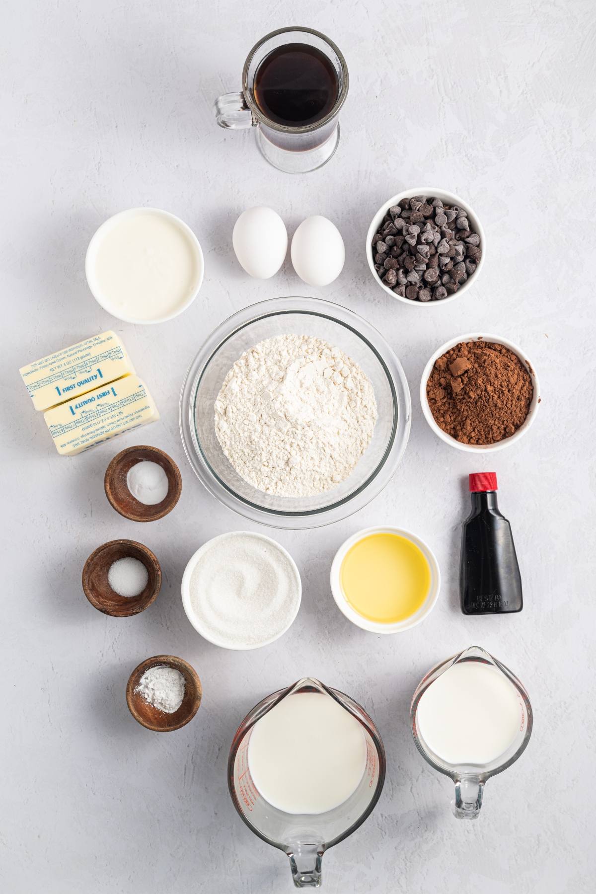 Baking ingredients arranged on a light surface, including flour, eggs, sugar, chocolate chips, cocoa, and milk.
