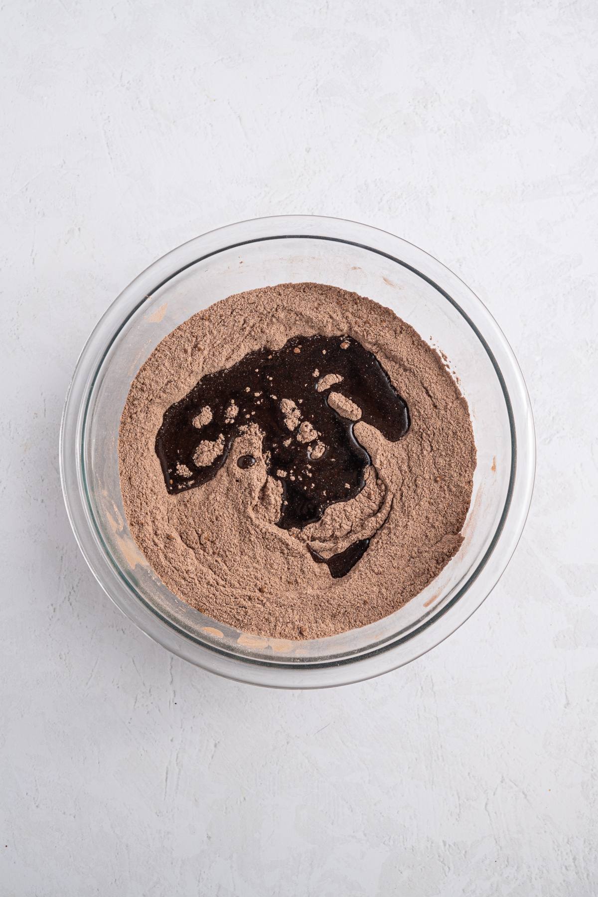 A glass bowl with cocoa powder mixture and liquid ingredients on a white surface.