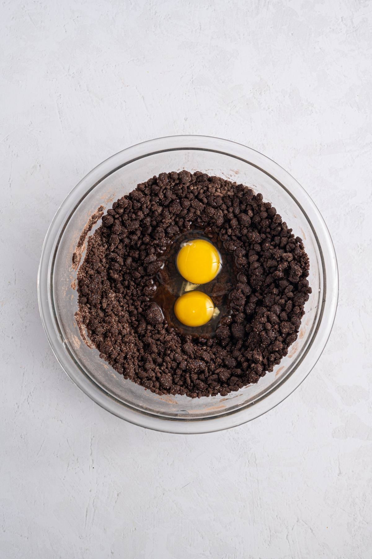 A glass bowl with chocolate crumb mixture and two raw eggs in the center, on a white surface.