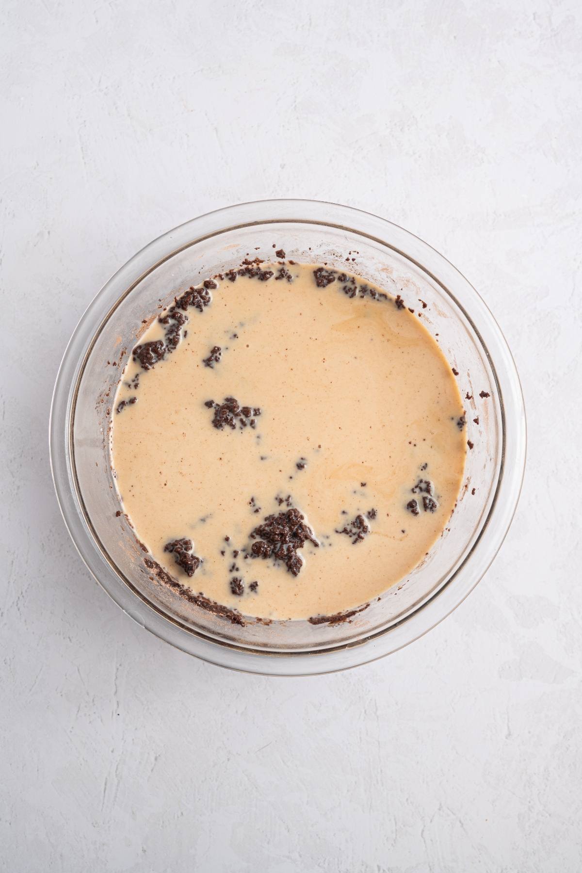 A glass bowl with a creamy batter and chunks of chocolate mixture on a light gray surface.