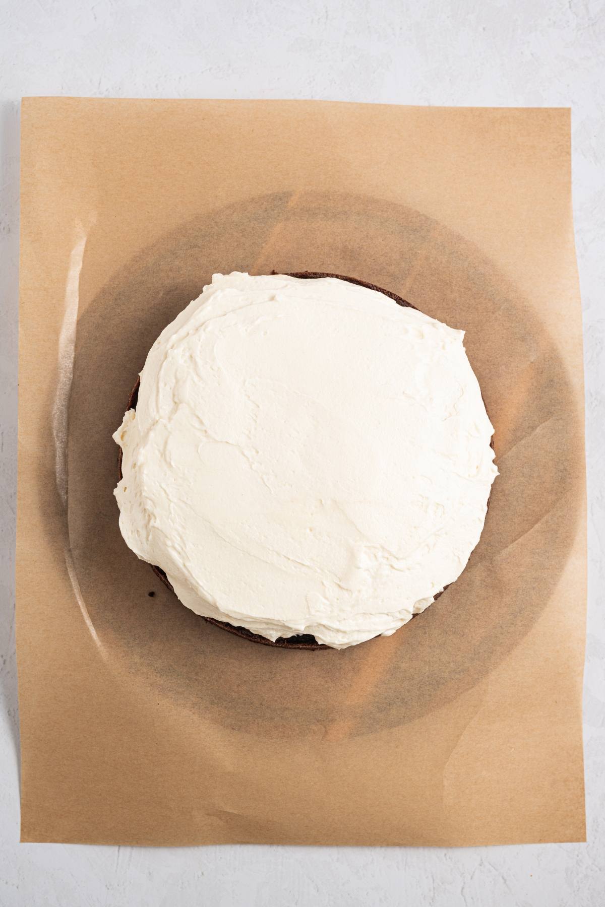 A round cake with white frosting on parchment paper, viewed from above.