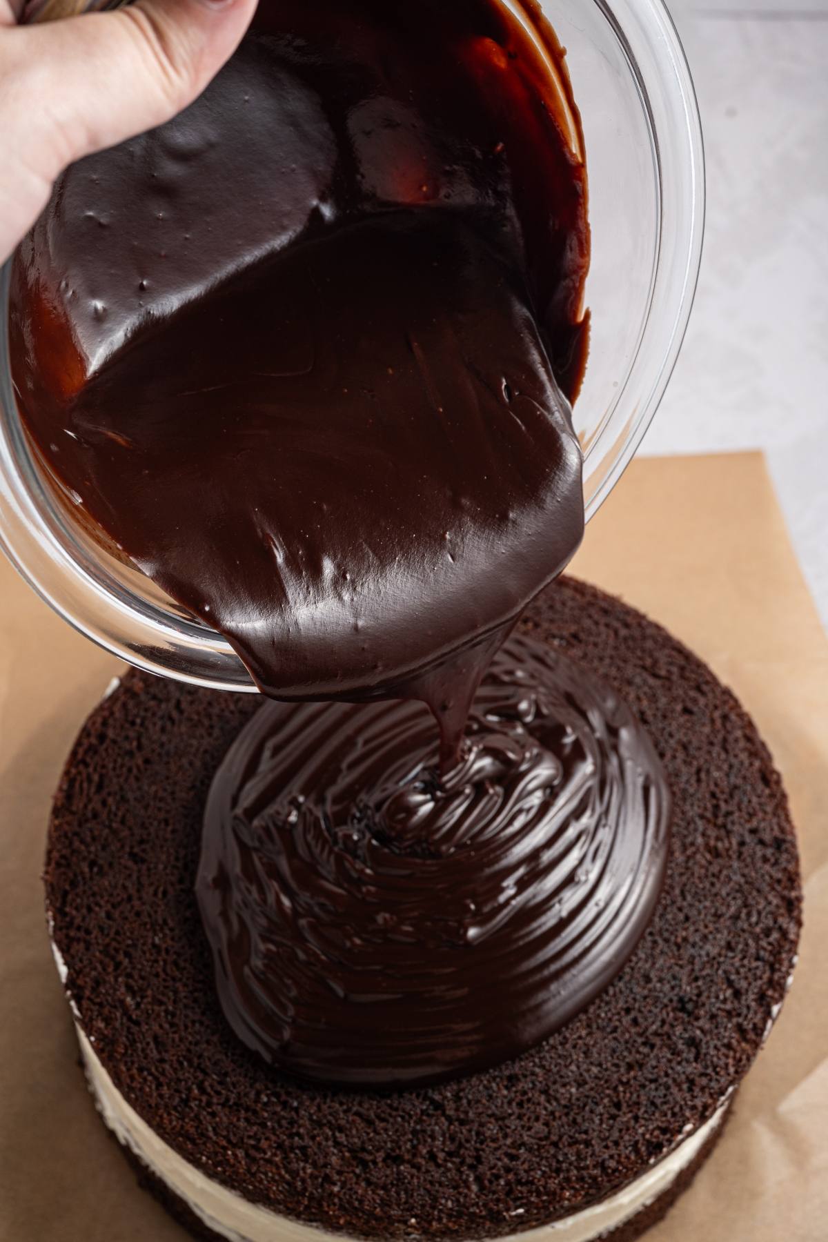 A bowl of chocolate ganache being poured onto a layered chocolate cake.