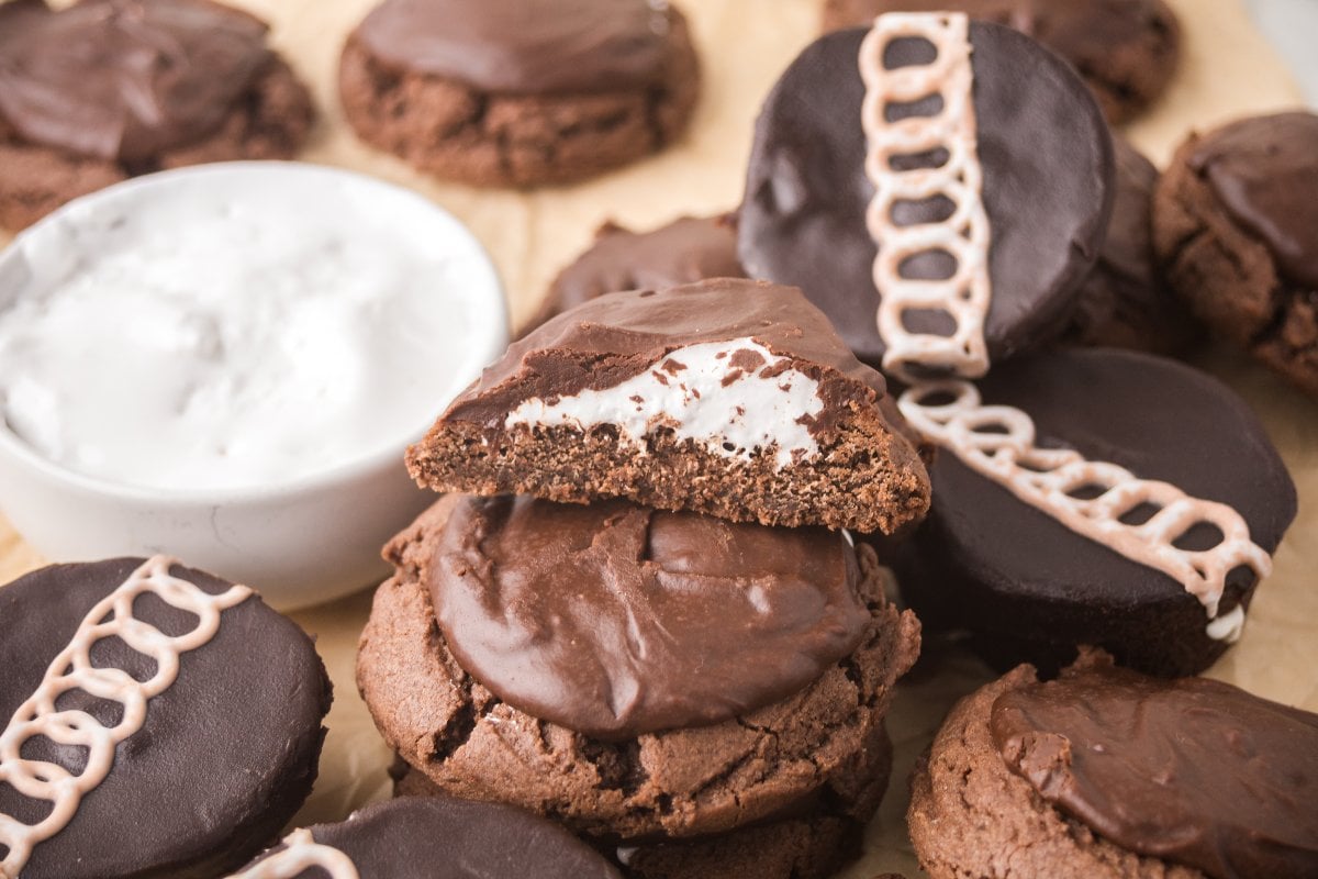 A stack of Ding Dong Cookies with cream filling, some topped with icing swirls, sits next to a bowl of white cream.