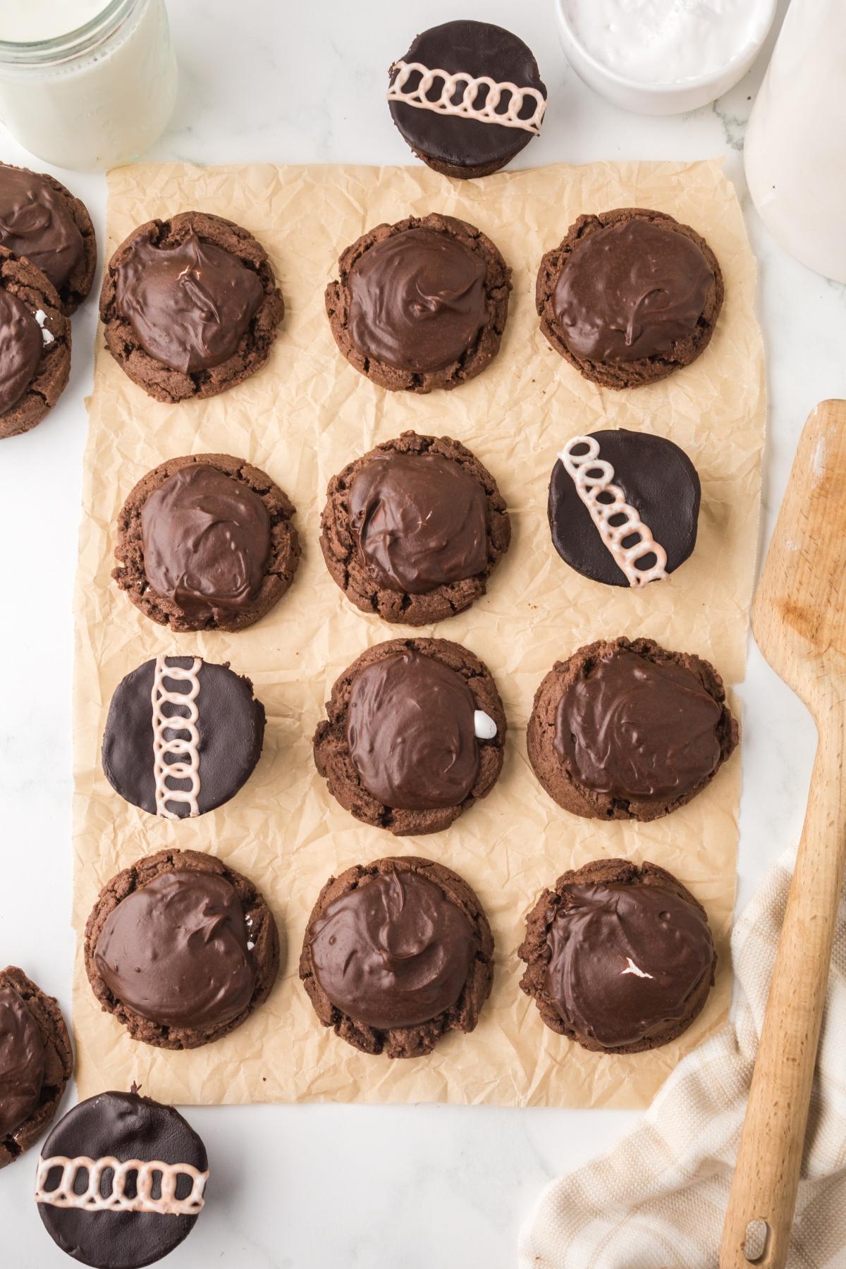 Chocolate cookies with glossy frosting on parchment, some decorated with white icing loops, wooden spoon beside them.