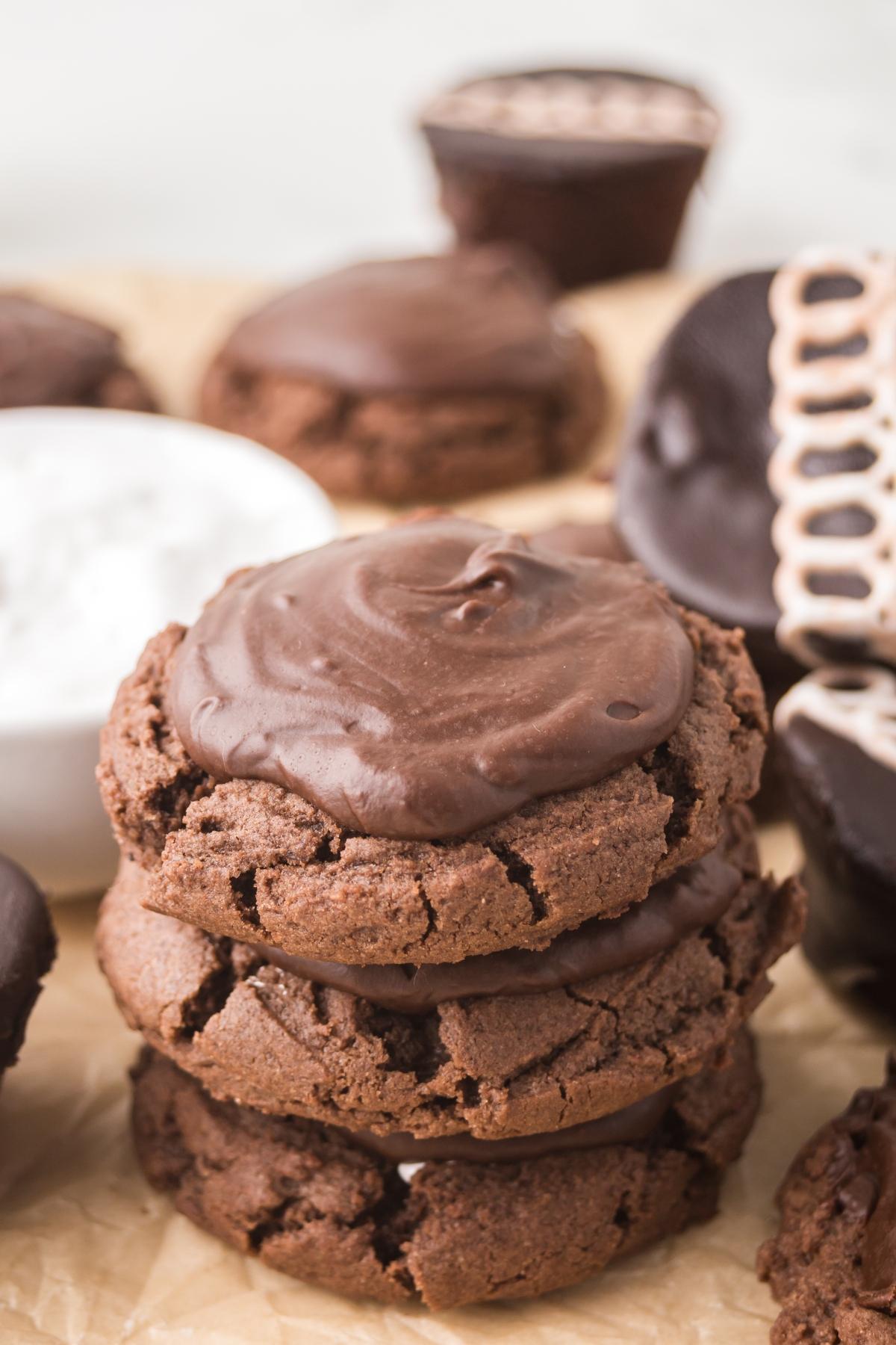 A stack of three frosted chocolate cookies surrounded by more cookies and a bowl of white icing.