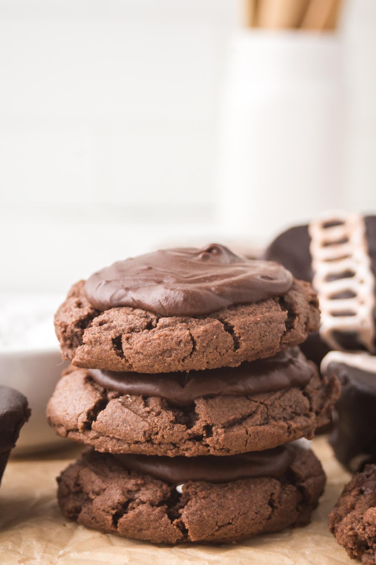 Three stacked chocolate cookies with chocolate frosting on parchment paper, with blurred kitchen items in the background.