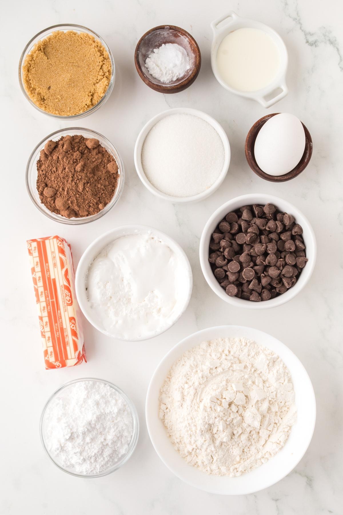 Top view of baking ingredients in bowls: flour, sugar, brown sugar, cocoa, chocolate chips, egg, milk, and butter.