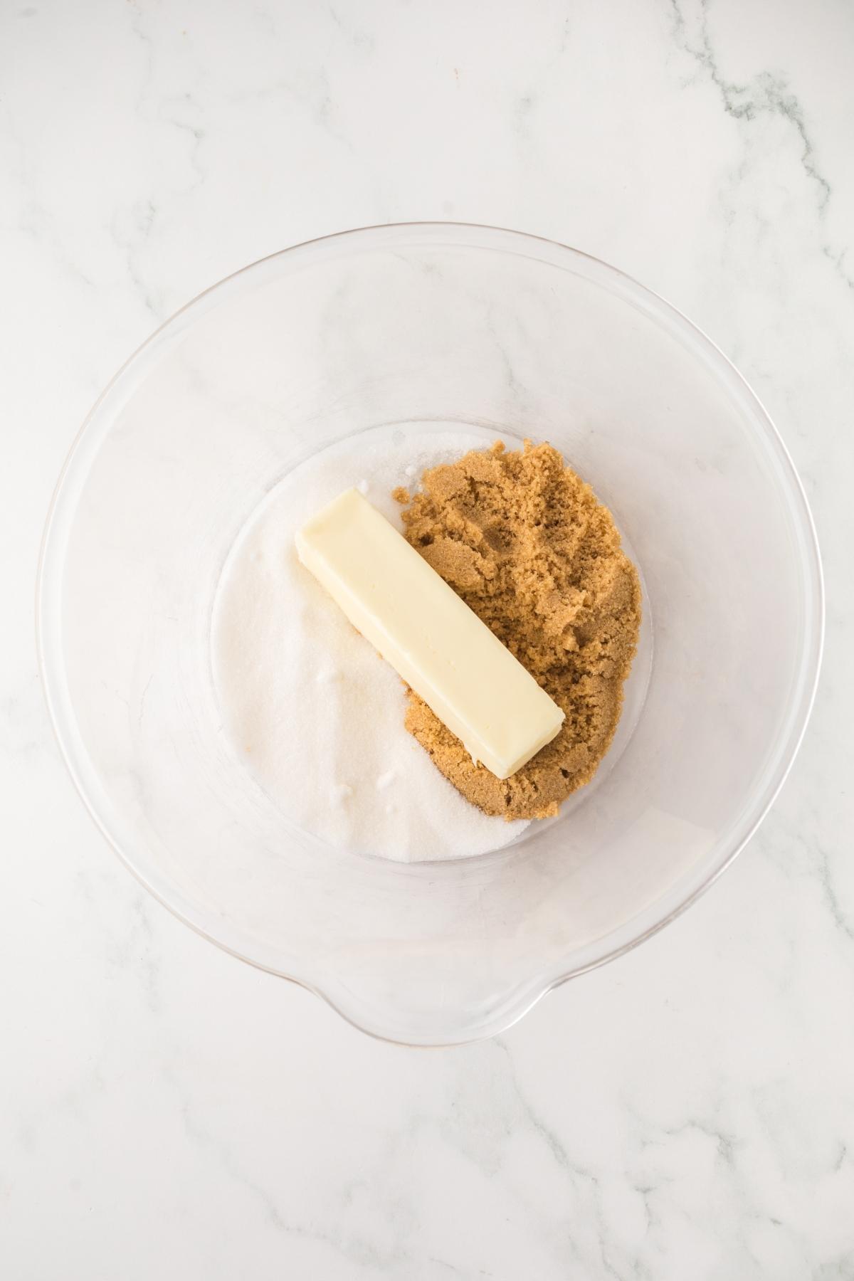 A mixing bowl with white sugar, brown sugar, and a stick of butter on a white marble surface.