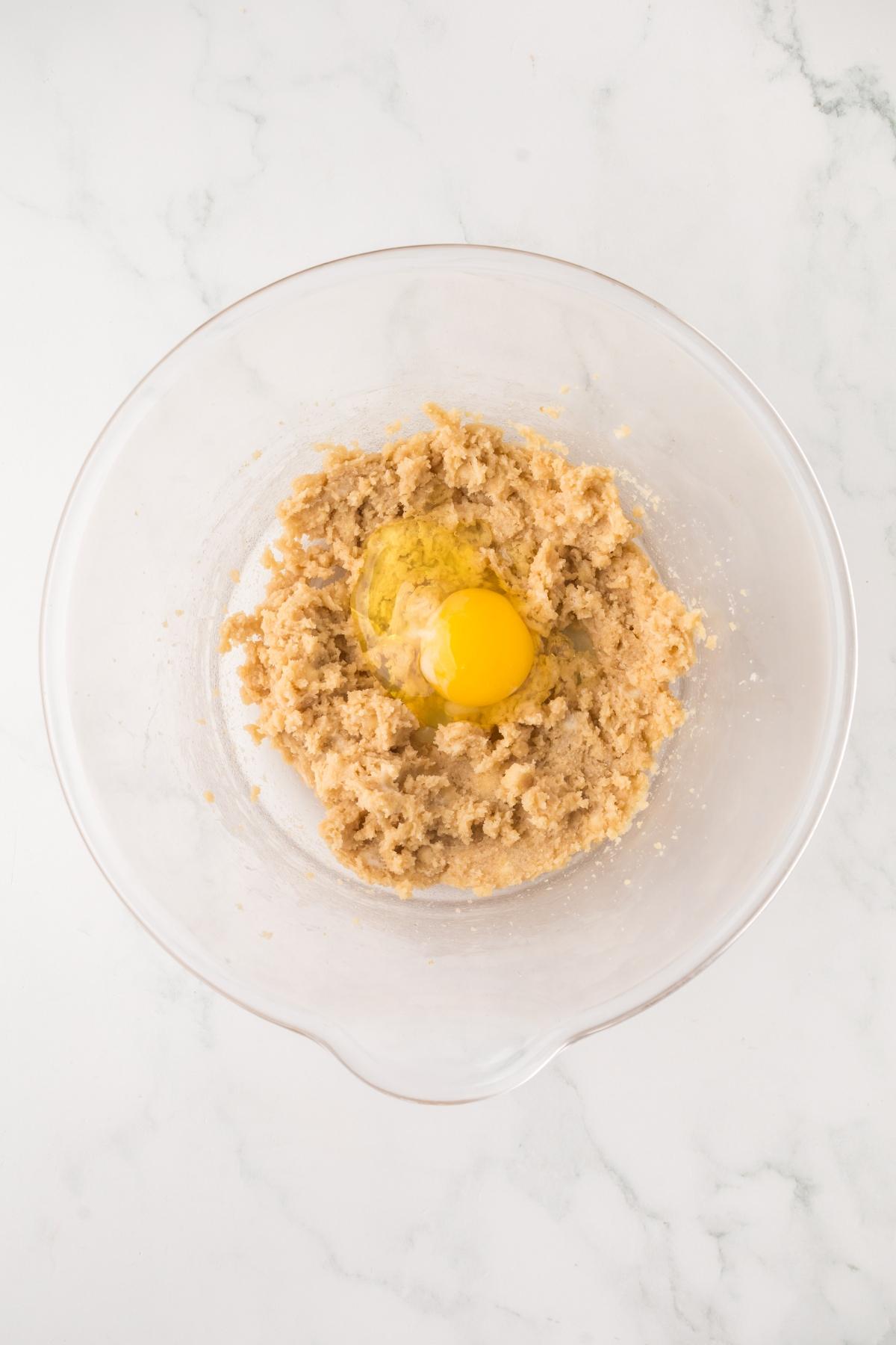 Clear glass mixing bowl with dough and a cracked raw egg in the center, on a white marble surface.