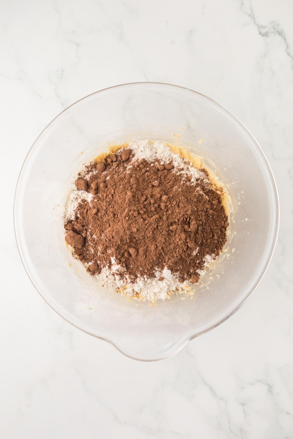 Clear glass mixing bowl with cocoa powder and flour on top of cookie dough batter on a white surface.