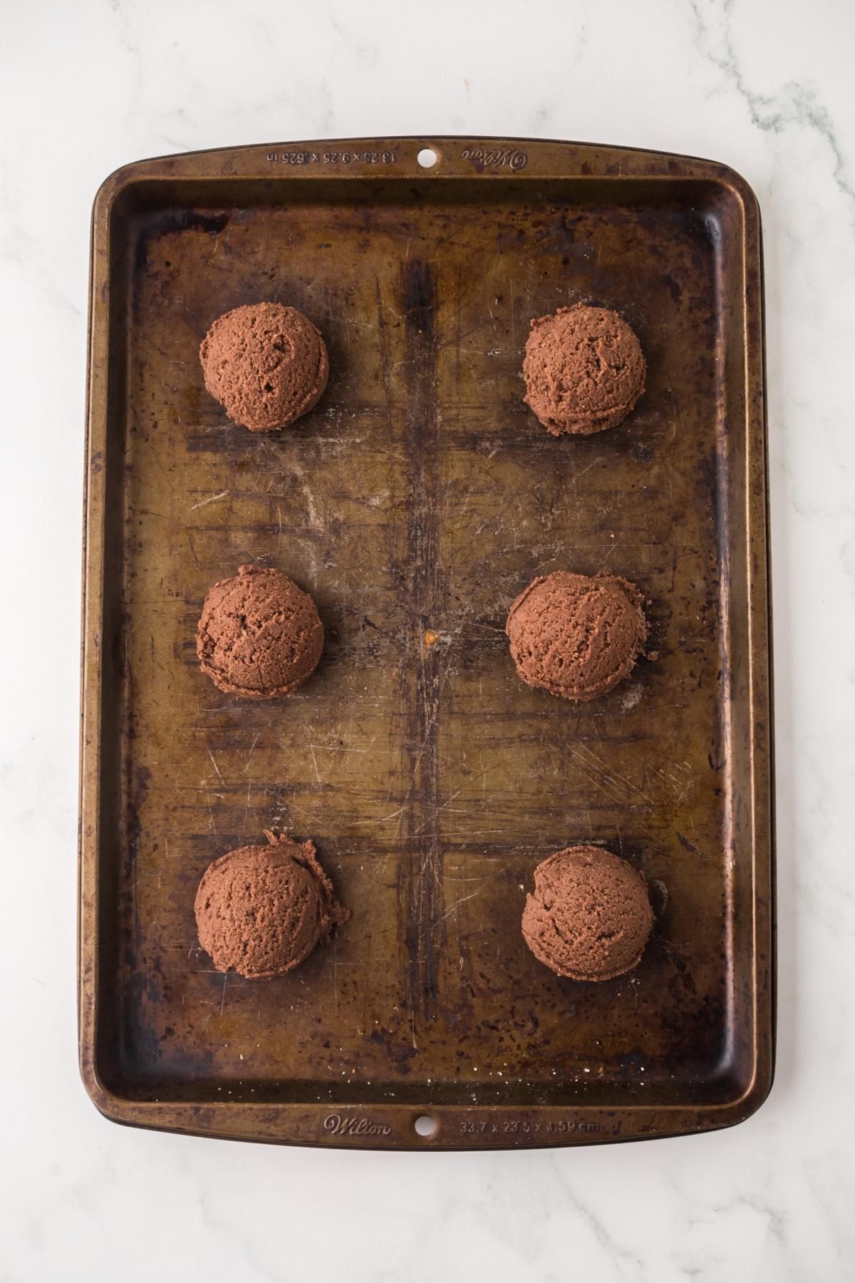 A baking sheet with six evenly spaced scoops of chocolate cookie dough on a white marble surface.