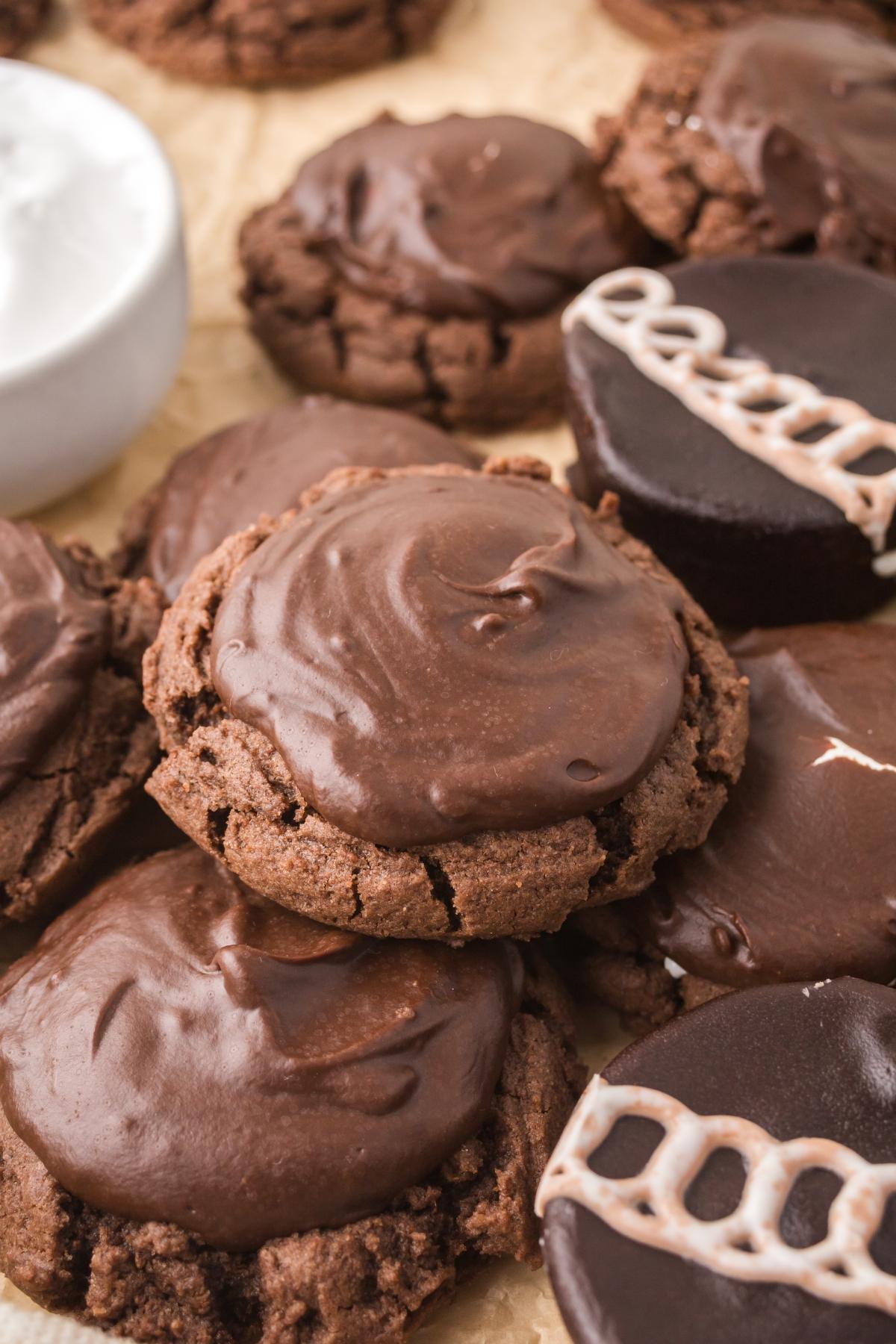 Chocolate cookies with rich chocolate frosting, some with white icing swirls, piled together on parchment paper.