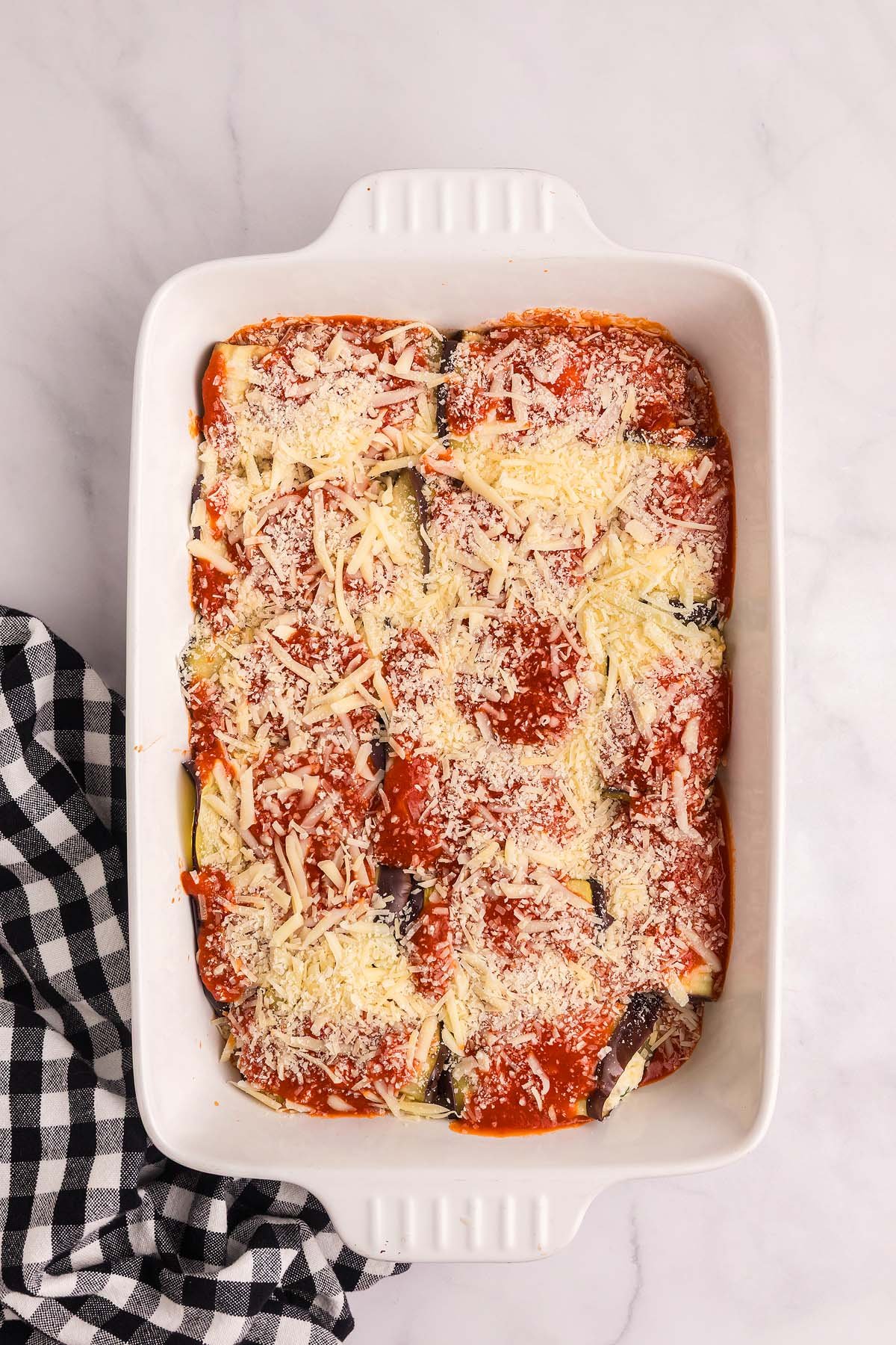 White baking dish with eggplant rollatini rolled eggplant slices topped with tomato sauce and shredded cheese—on a marble surface.