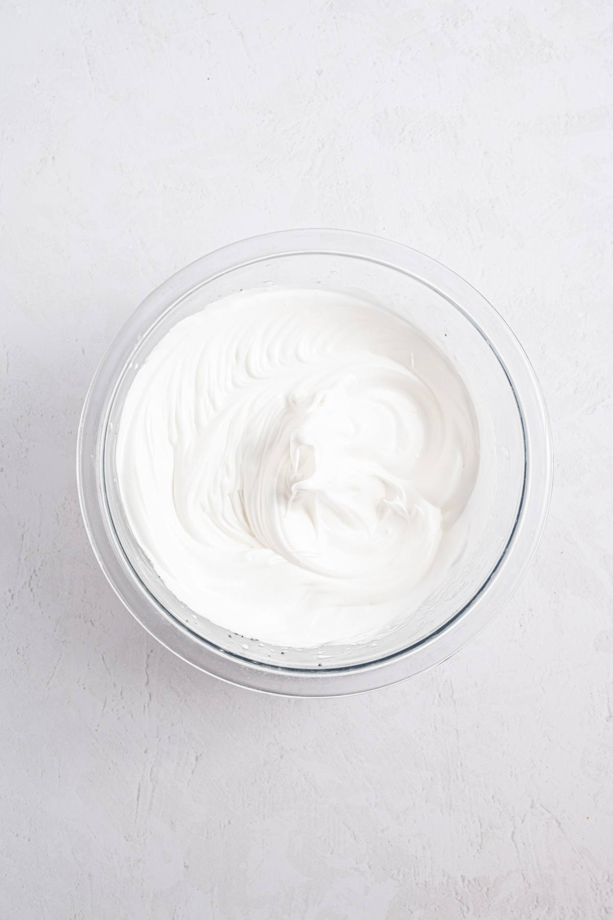 A glass bowl filled with fluffy whipped cream on a light textured surface.