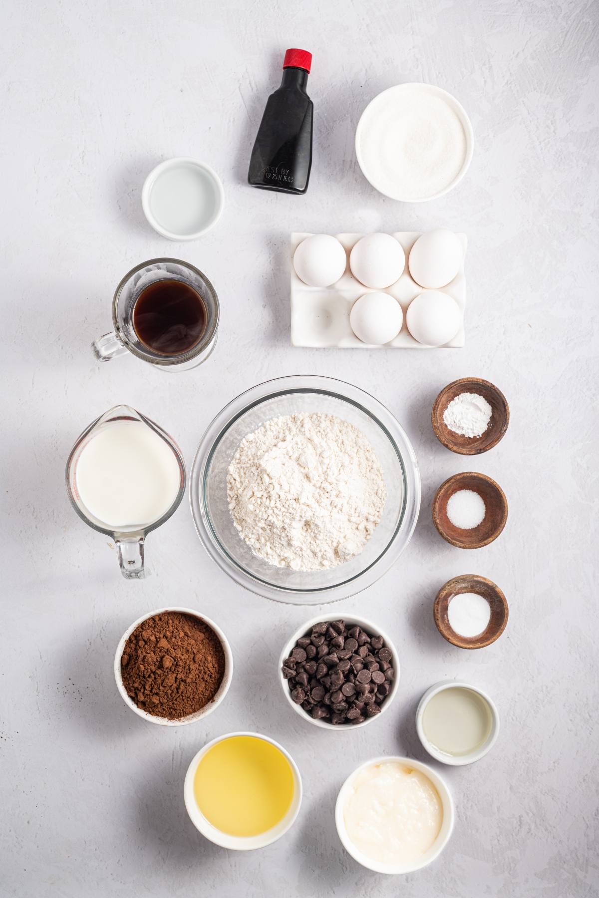 Baking ingredients arranged neatly on a white surface, including eggs, flour, cocoa, sugar, and chocolate chips.