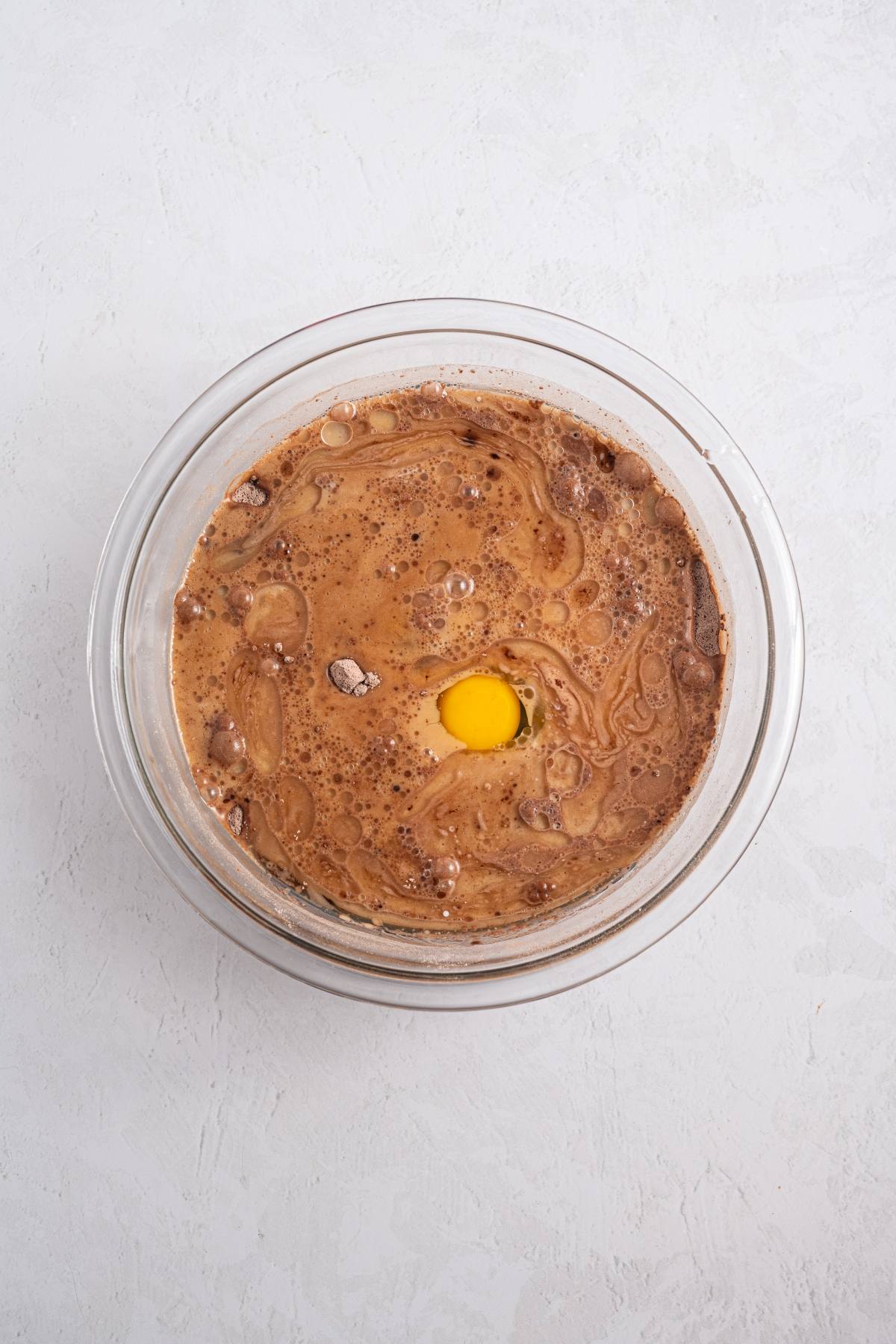 A glass bowl with chocolate cake batter and a raw egg on a white surface.