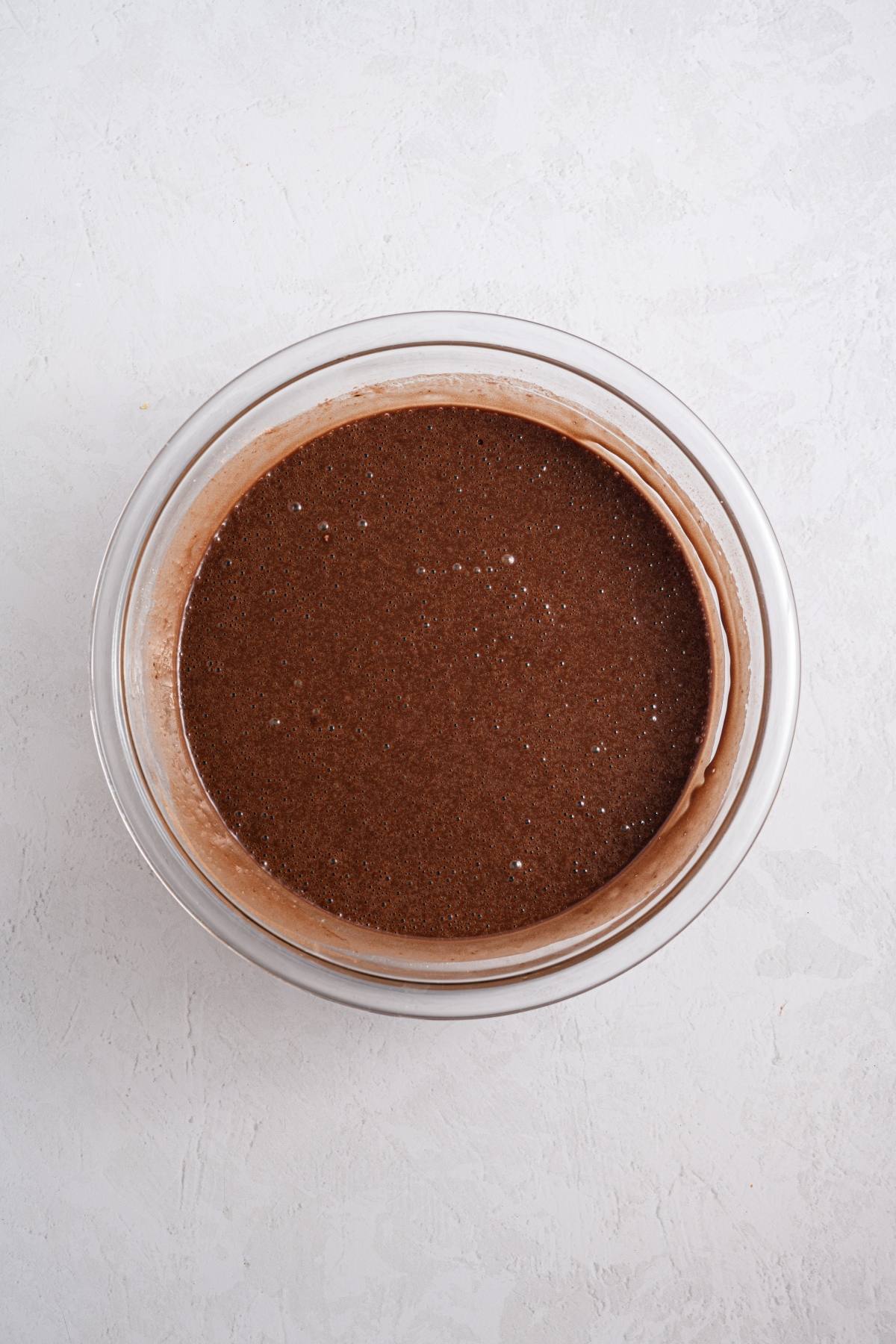 A glass bowl filled with chocolate cake batter on a light, textured surface.