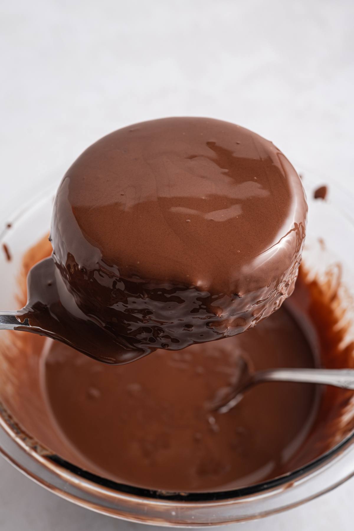 A fork holds a round cake coated in glossy chocolate above a bowl of melted chocolate.