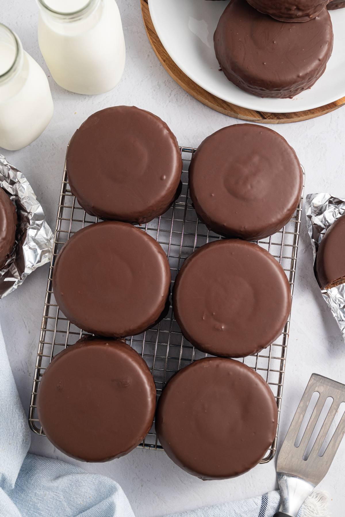 Six round chocolate-covered cakes on a cooling rack with bottles of milk and a spatula nearby.