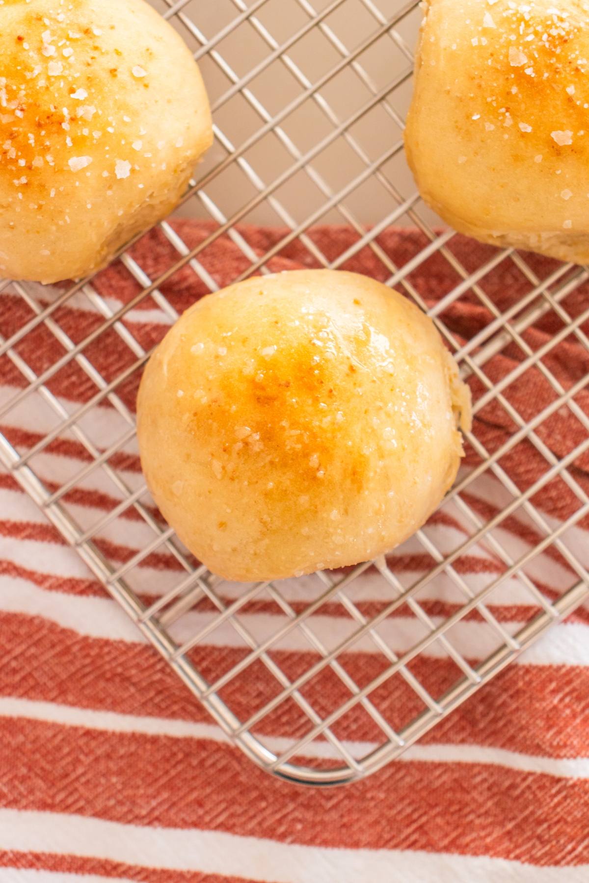 Three golden brown bread rolls with sprinkled salt on a cooling rack over a red and white striped towel.
