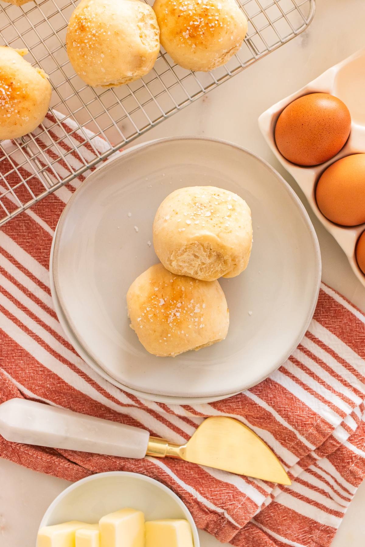 Two dinner rolls on a plate, surrounded by eggs, butter, and more rolls on a cooling rack.