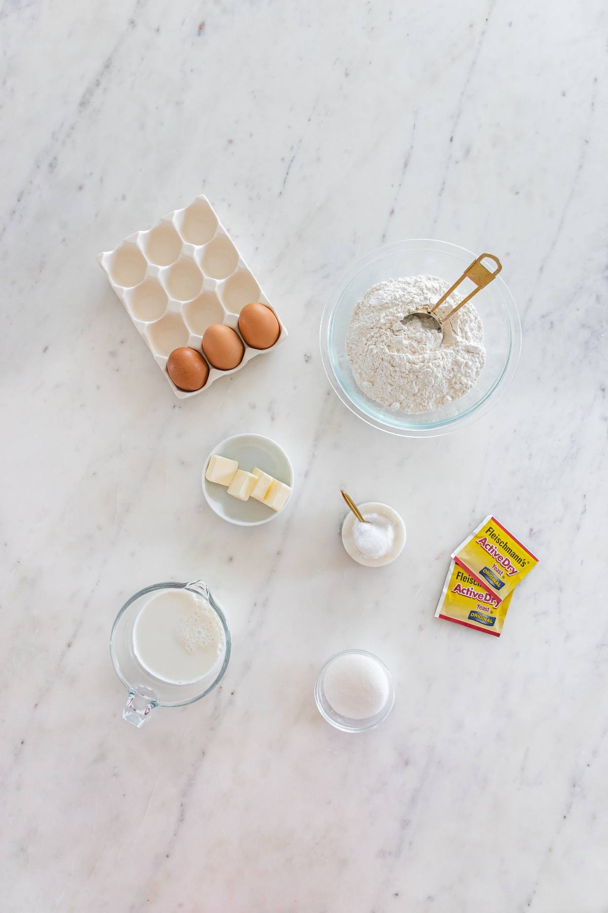 Baking ingredients on a marble countertop: eggs, flour, butter, salt, sugar, milk, and yeast packets.