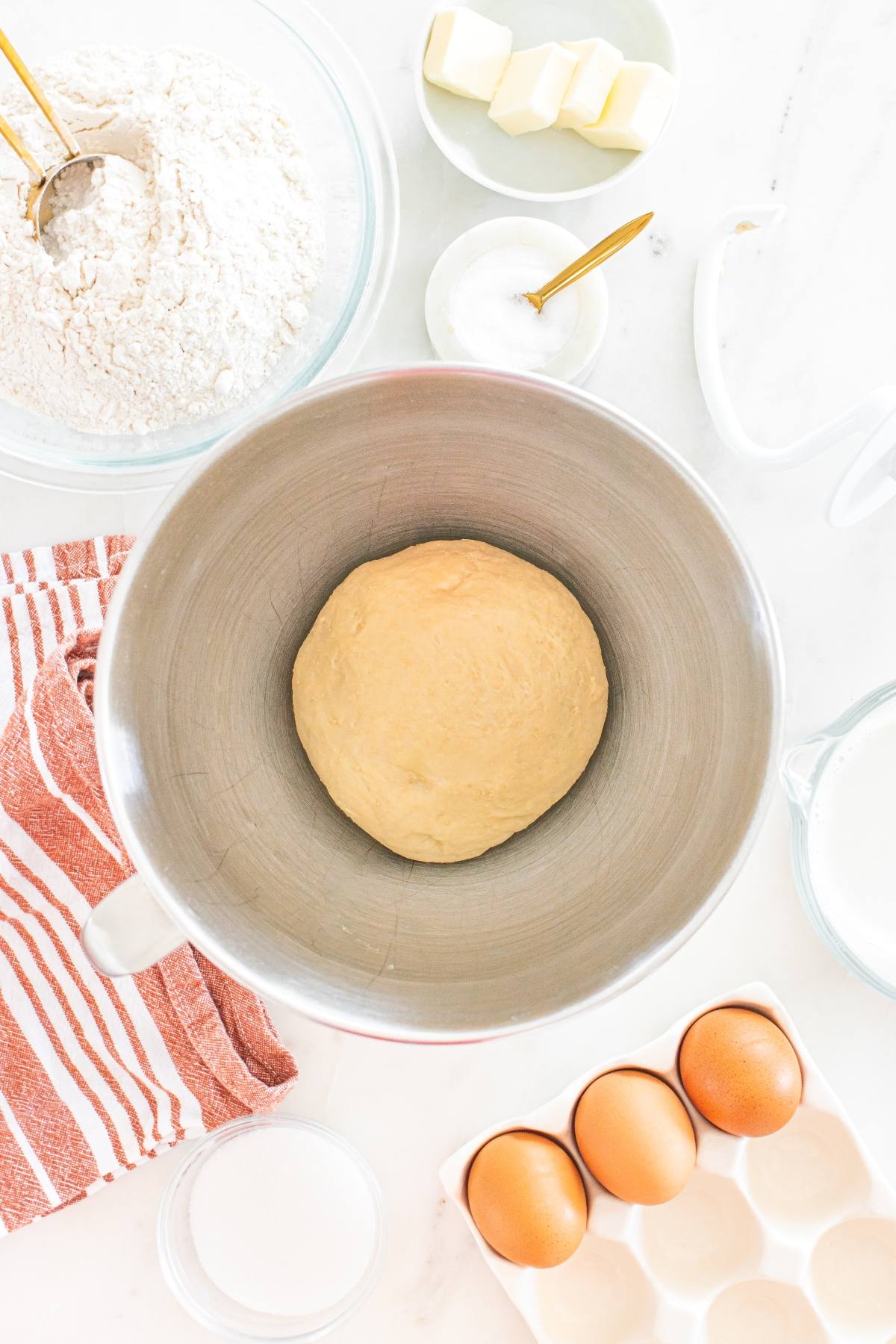 Ball of dough in a mixing bowl with flour, sugar, butter, eggs, milk, and a striped towel on a white surface.